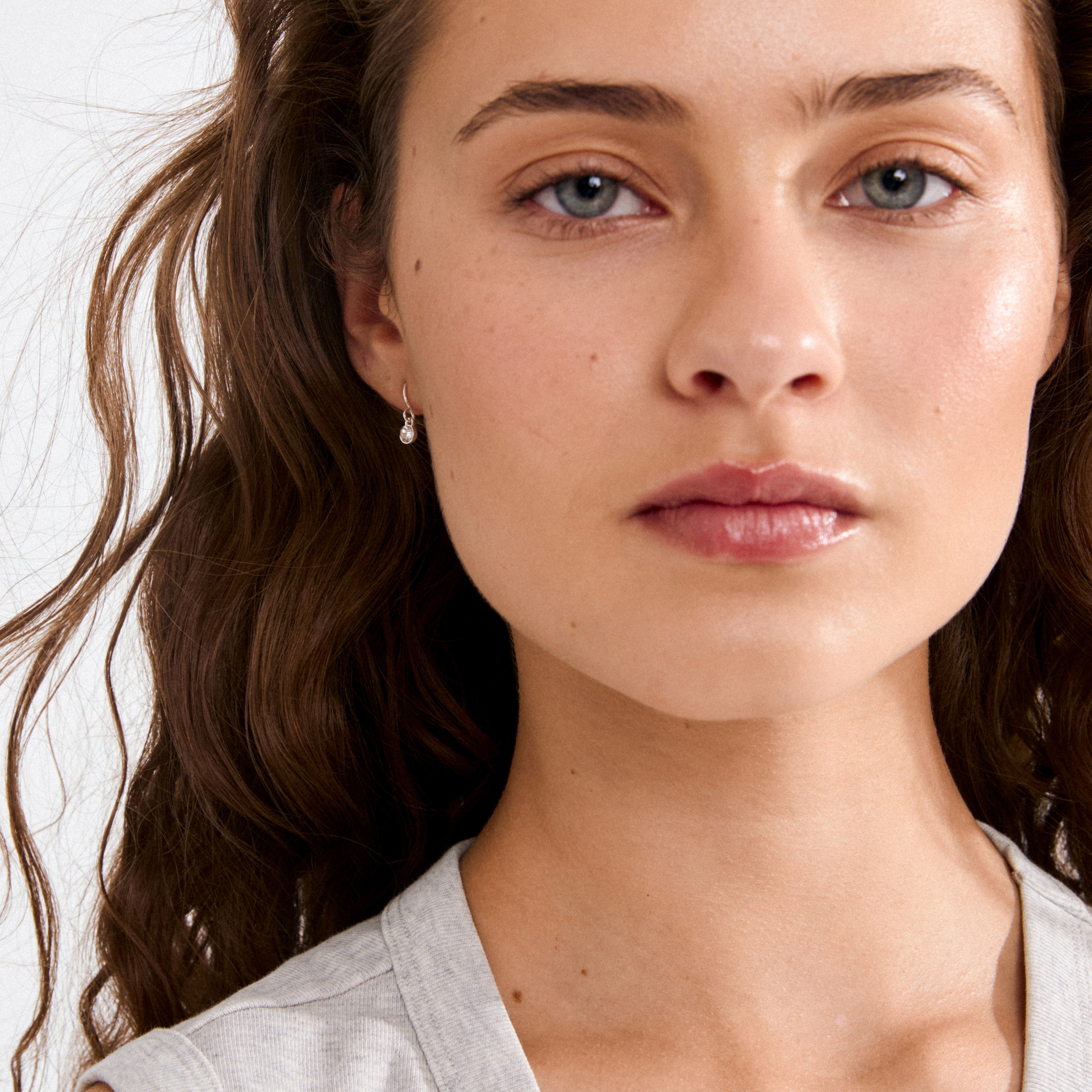 Young woman wearing small silver LUCIA crystal earstuds, showcasing their subtle sparkle against her wavy brown hair and simple grey top.