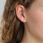 Woman's ear adorned with CLARA crystal cross earrings, silver-plated, featuring sparkling crystals for a feminine touch.