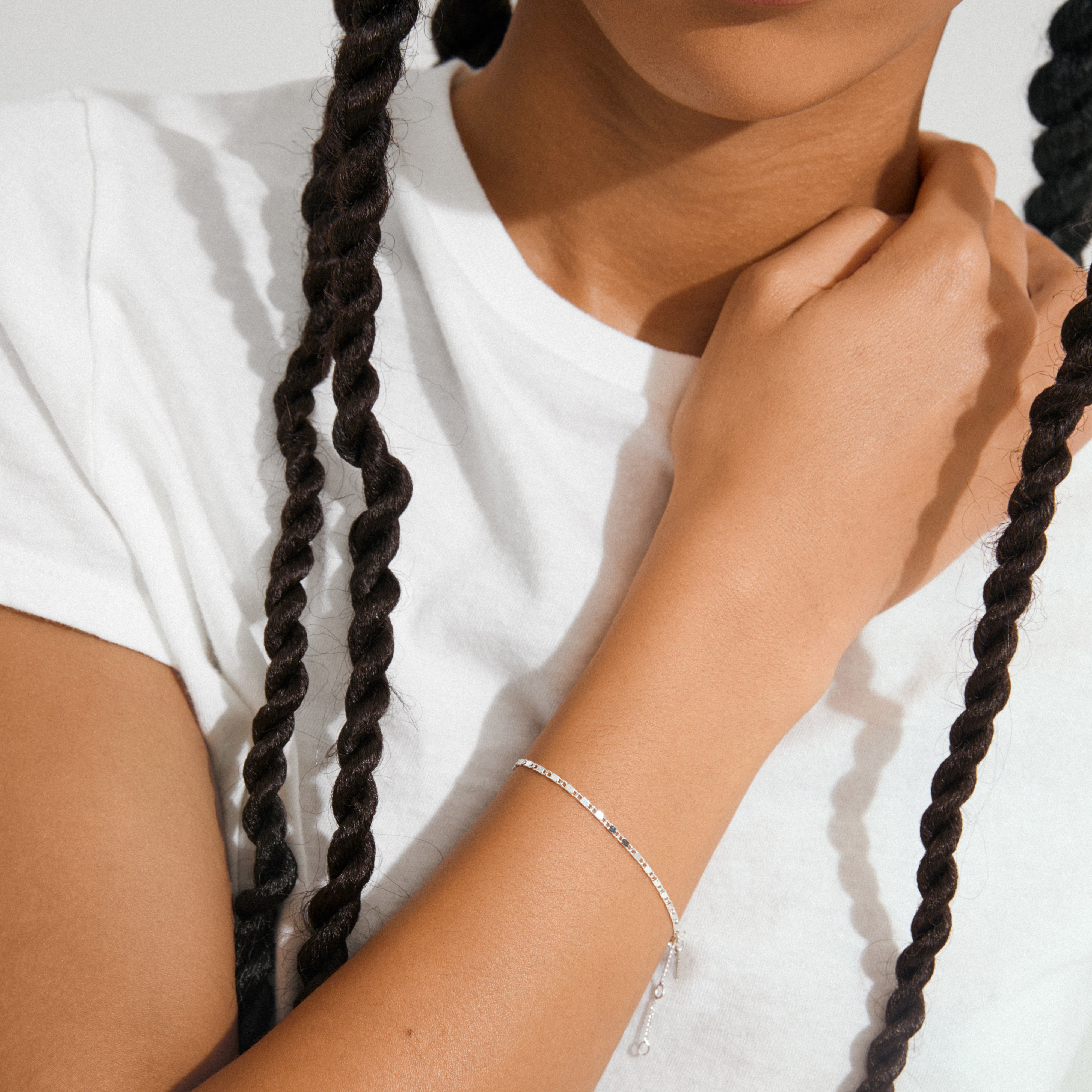 Person wearing a white T-shirt and PARISA flat link chain bracelet, silver-plated, gently touching their neck, showcasing the bracelet's delicate texture and reflective quality.