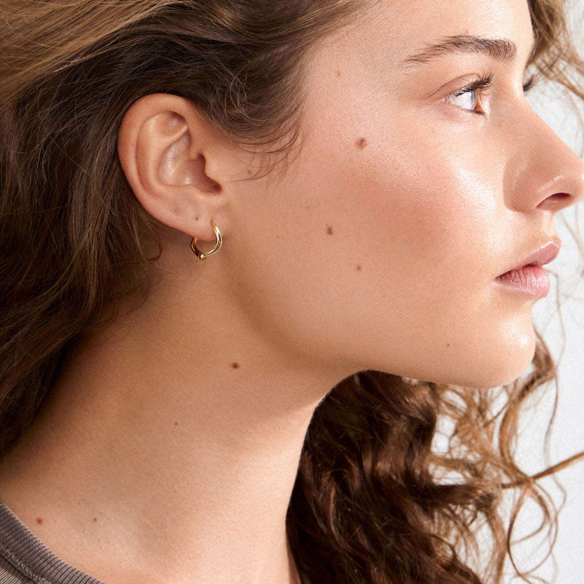 Young woman with brown hair wearing Alberte gold-plated hoop earring, showcasing its organic, sculptural design inspired by nature's shapes.