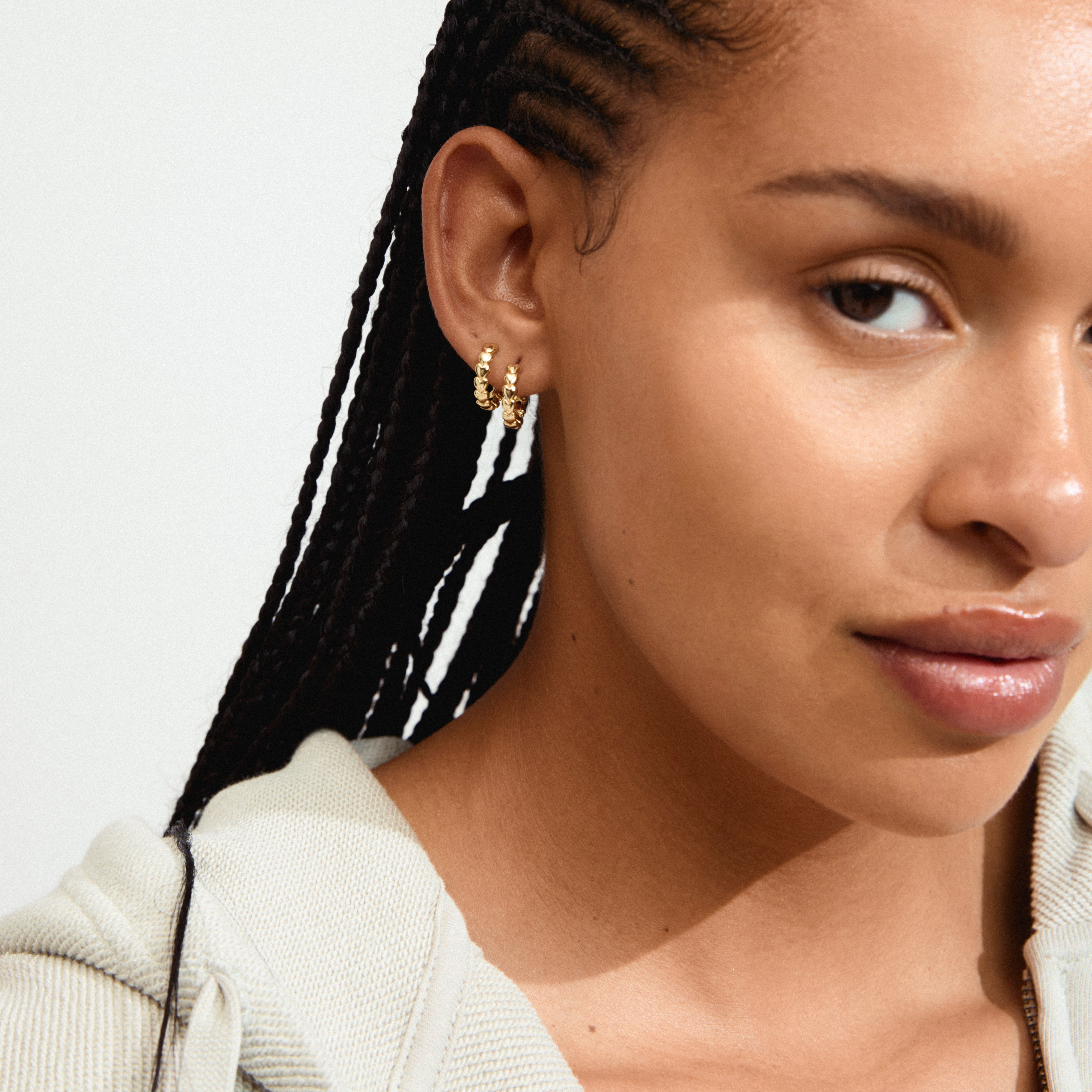 MAEVE hoop earrings, gold-plated with heart design, worn by a woman with braided hair. Perfect for a chic, versatile style.