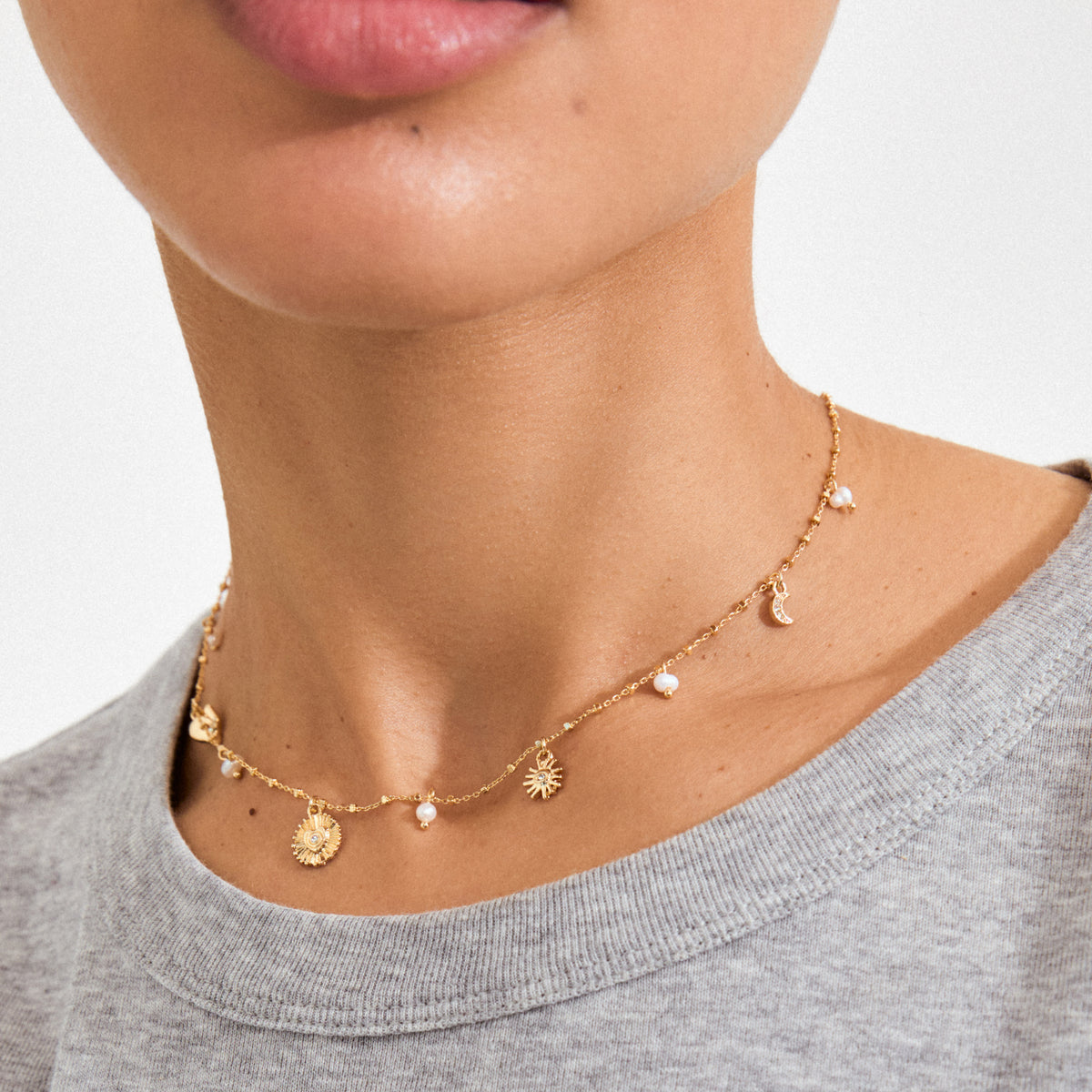 Close-up of MYLAN necklace, gold-plated with sunburst, crescent moon charms, and tiny pearls, worn over a light grey top.