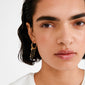 Close-up of MYLAN gold-plated hoop earrings with dangling chains and charms, including beads and a heart, worn by a woman with short dark hair.