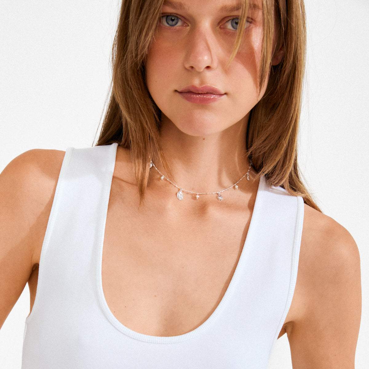 Young woman wearing the MYLAN necklace, silver-plated with sun, moon, and heart charms, highlighting its delicate, whimsical design above a simple white tank top.