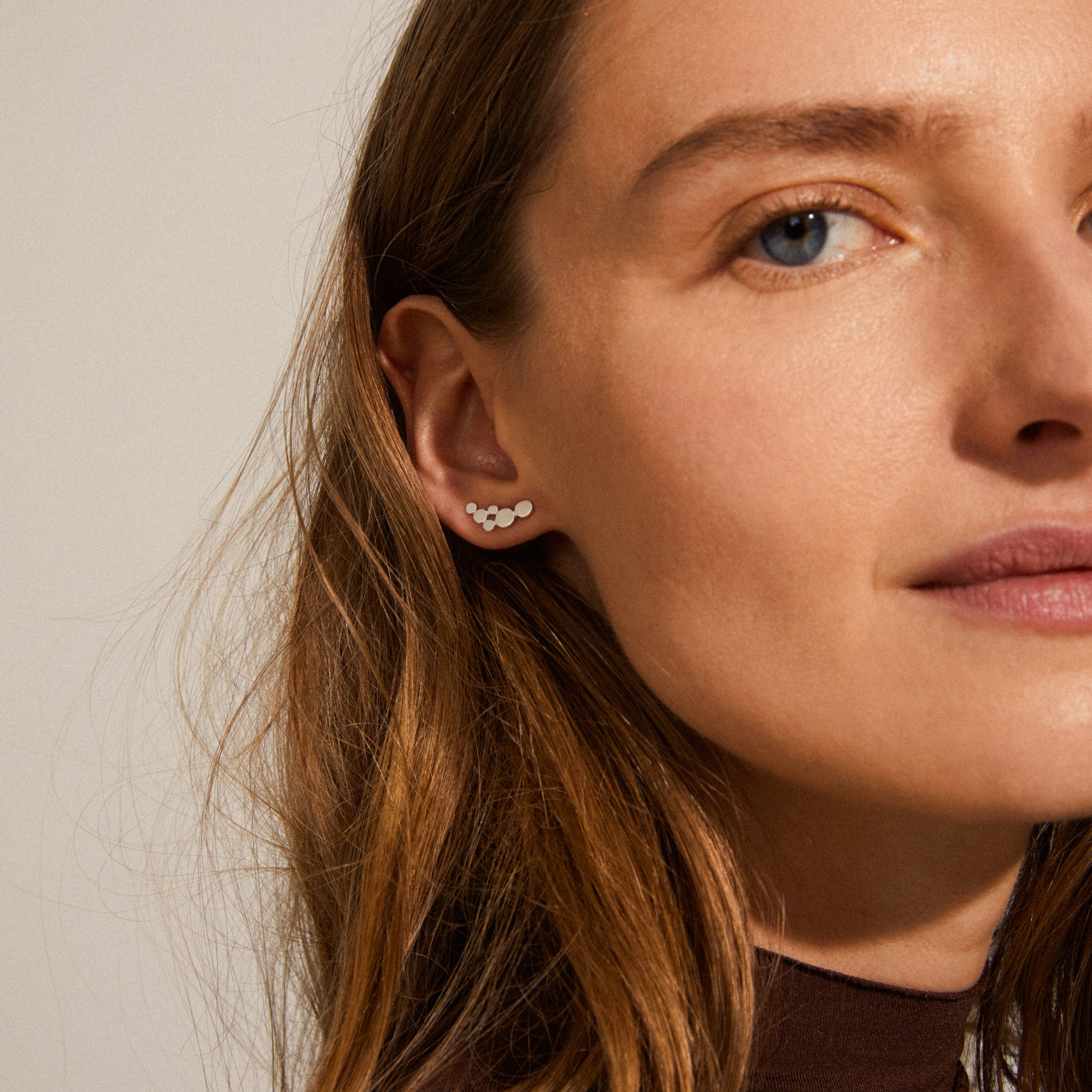 Close-up of a woman's ear showcasing the LEAH earring, a silver-plated, satin-matte stud with small circles, embodying timeless elegance and Scandinavian craftsmanship.