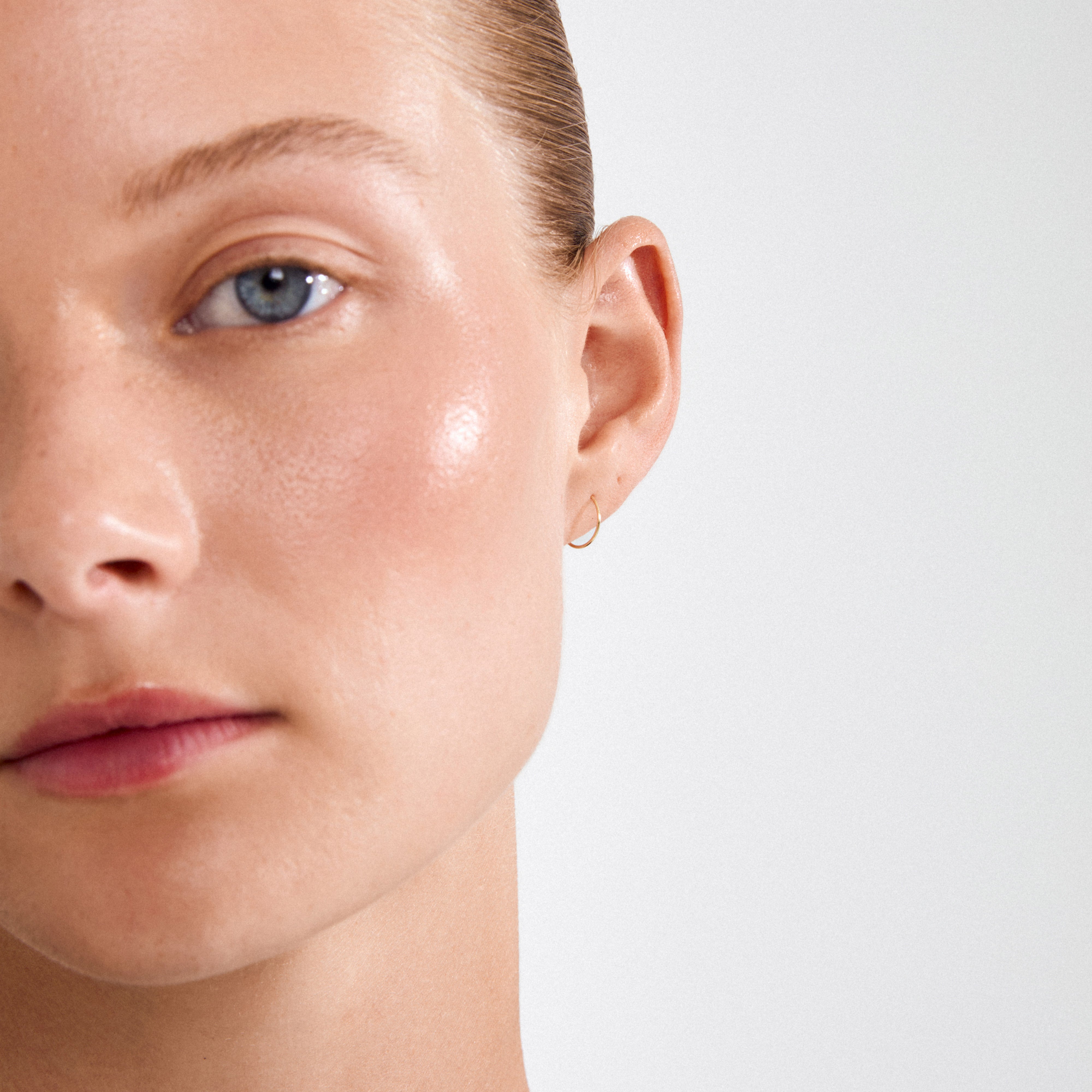 Close-up of a person wearing RAQUEL mini hoop earrings gold-plated, showcasing smooth, dewy skin and a blue-gray eye, highlighting the earring's elegant, timeless design.