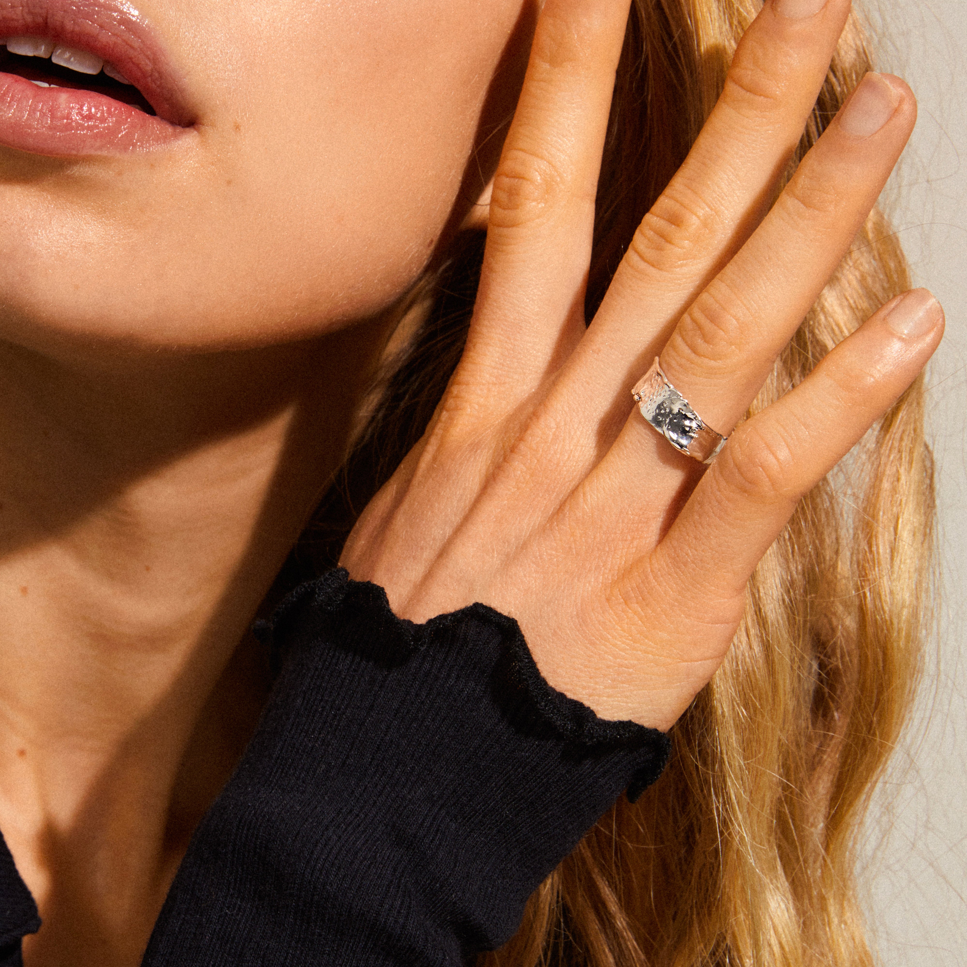 Woman wearing the BATHILDA rustic ring, a silver-plated, hammered-texture piece on her middle finger, showcasing bold elegance against a black ruffled-edge sleeve.
