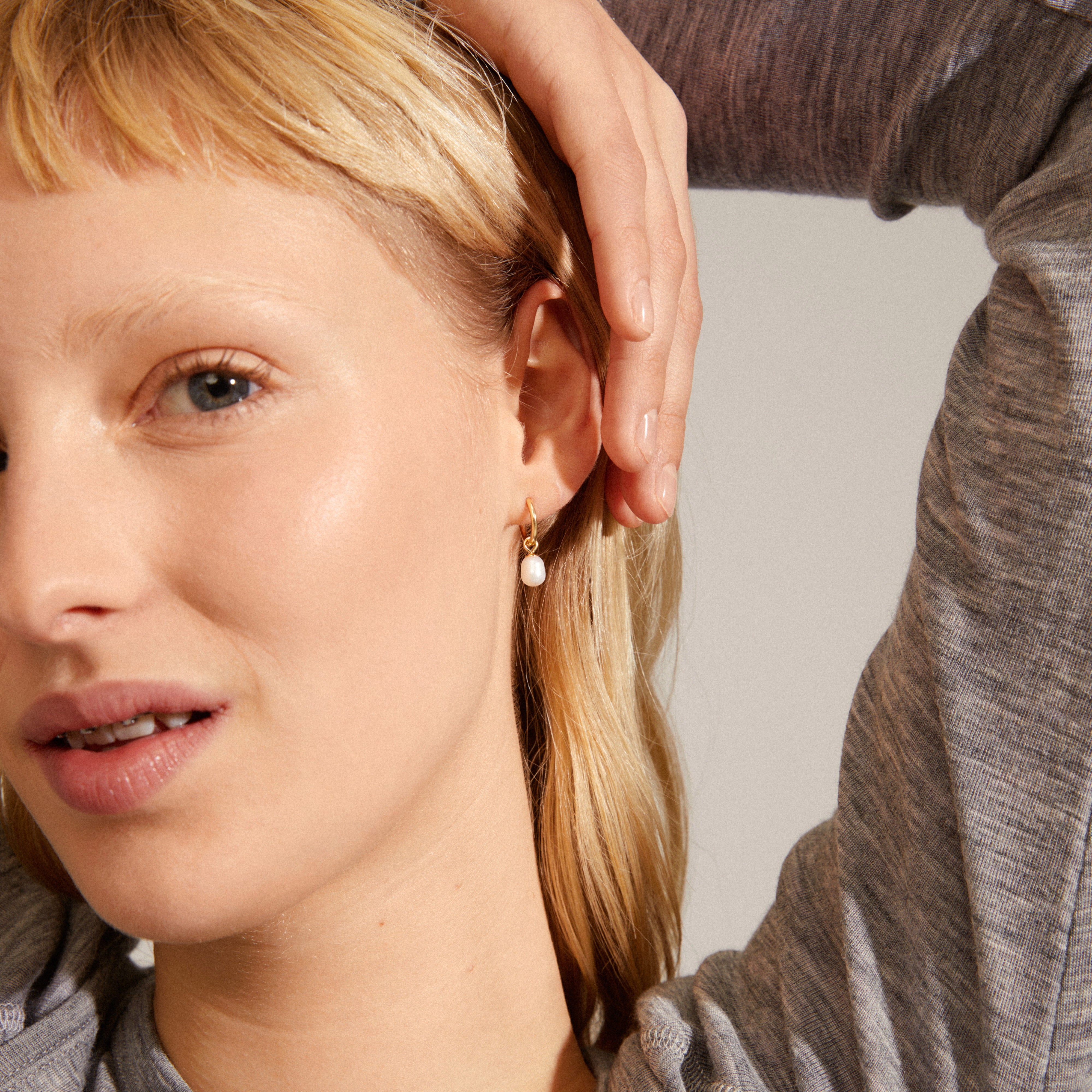 BERTHE pearl hoop earrings gold-plated; woman showcasing the earrings with her hand near ear, highlighting their elegant, natural design with white freshwater pearl pendants.