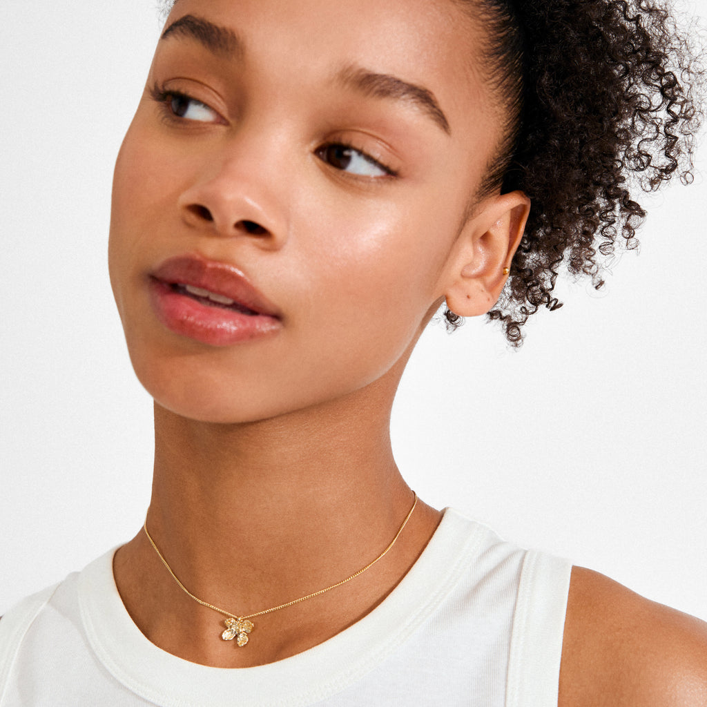 Close-up of AJANI gold-plated necklace by Pilgrim, featuring a delicate floral pendant on a young woman wearing a white top and small gold stud.