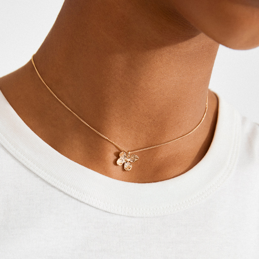 Close-up of the AJANI gold-plated necklace from Pilgrim, featuring a delicate textured floral pendant on a fine chain, worn above a white crew-neck top.