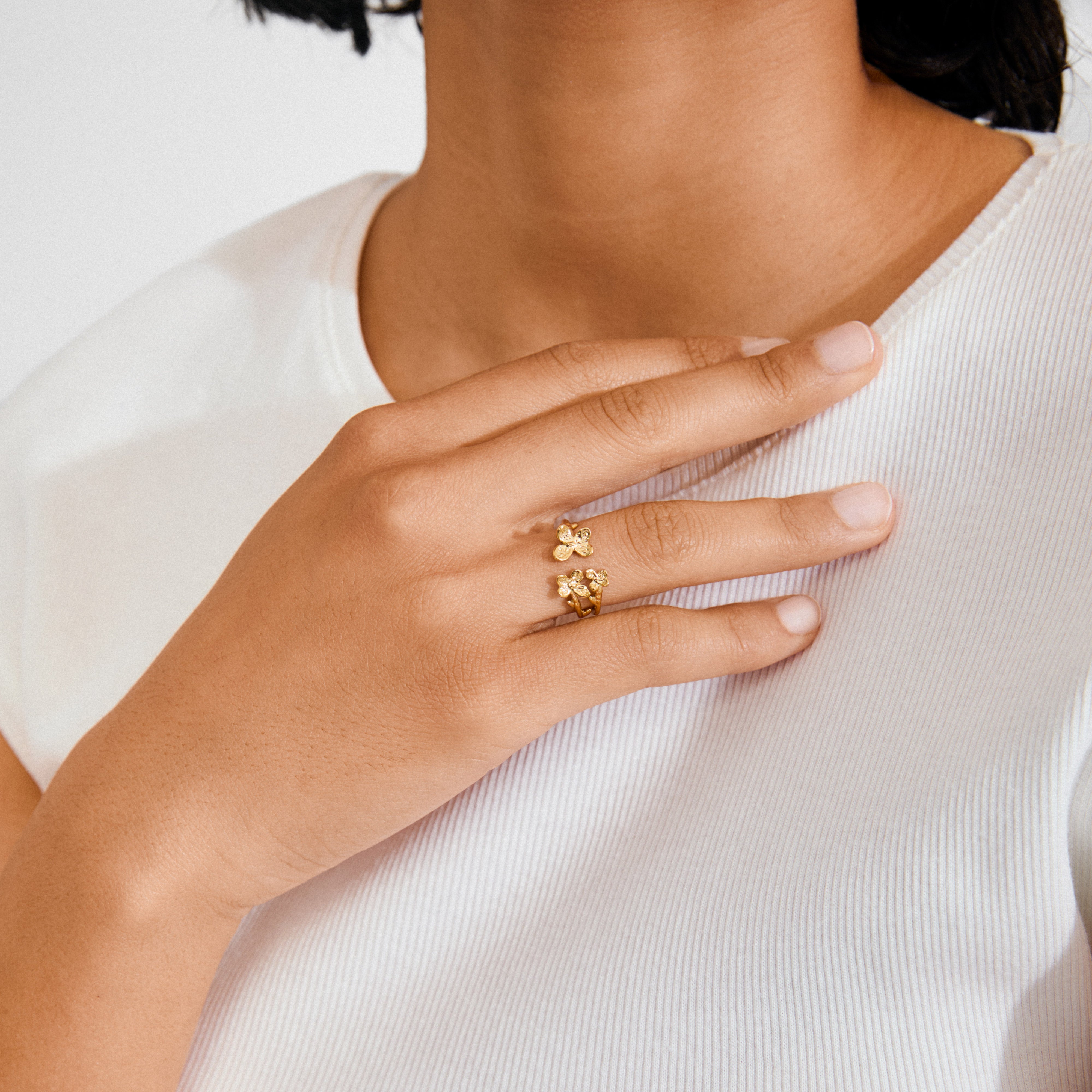 Close-up of a hand wearing Pilgrim's gold-plated AJANI rings with floral motifs on middle and ring fingers, against a white ribbed top.