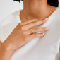 Close-up of a hand wearing Pilgrim's gold-plated AJANI rings with floral motifs on middle and ring fingers, against a white ribbed top.