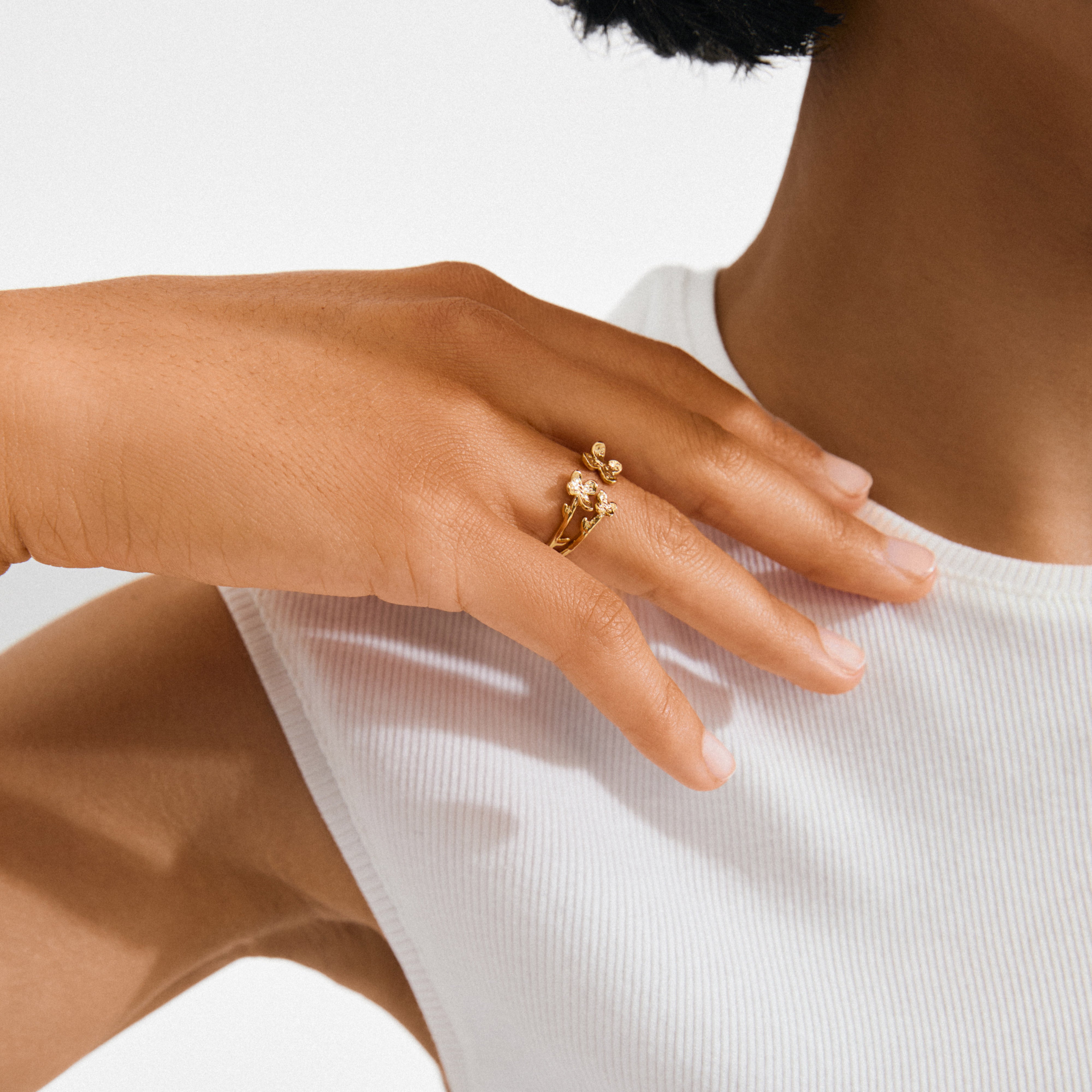 Hand wearing the AJANI gold-plated ring featuring twig-inspired flower motifs with gemstones, showcasing Pilgrim’s nature-inspired, handcrafted Danish design on a white ribbed top.