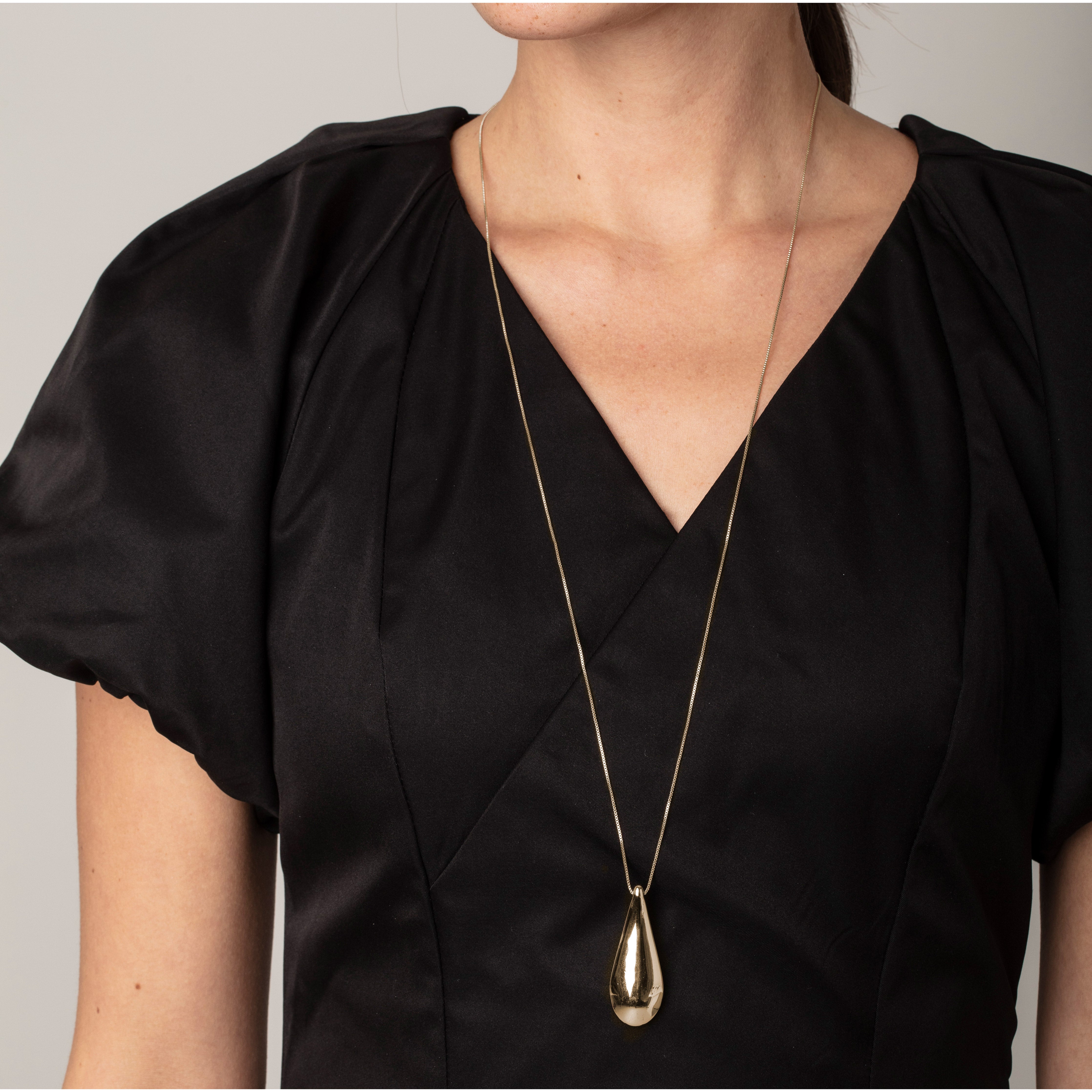 ALMA necklace gold-plated featuring a large teardrop pendant with mirror and satin finishes, worn by a woman in a black dress.