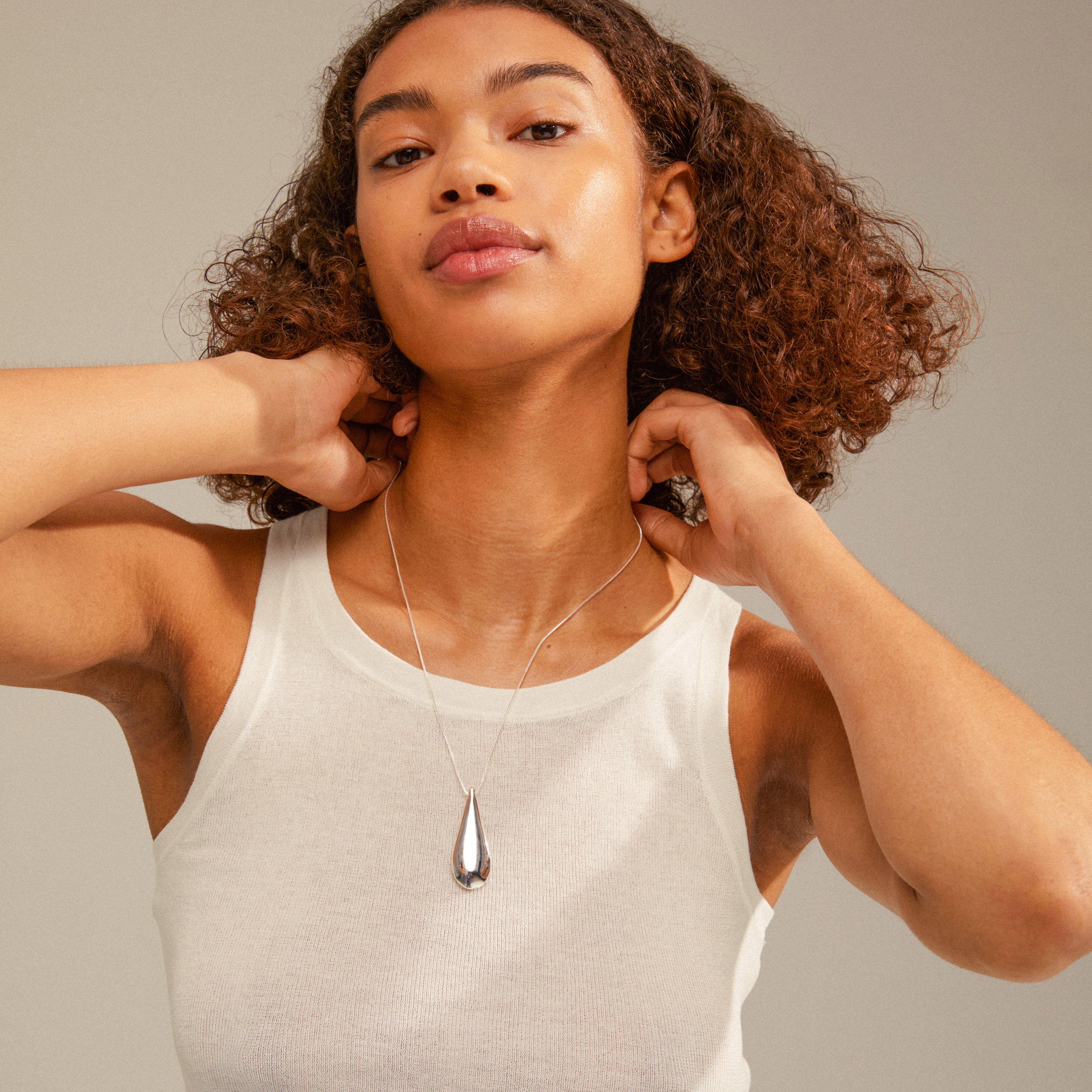 Woman wearing ALMA necklace; silver-plated, adjustable teardrop pendant with mirror and satin finishes, complementing her white tank top and curly hair.