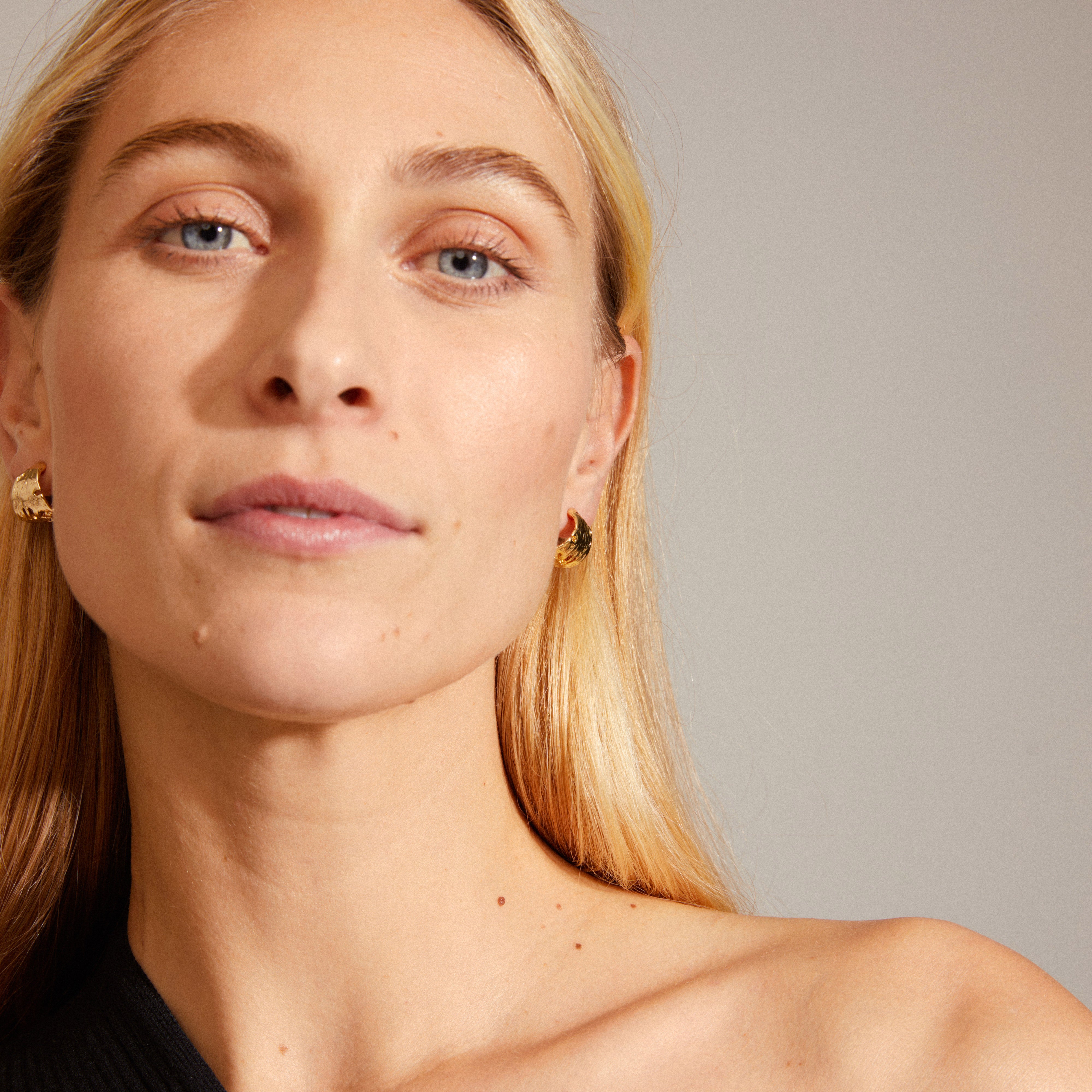 Close-up of a woman wearing BRENDA gold-plated earrings by Pilgrim, showcasing their dynamic, wide semi-hoops with irregular surfaces.
