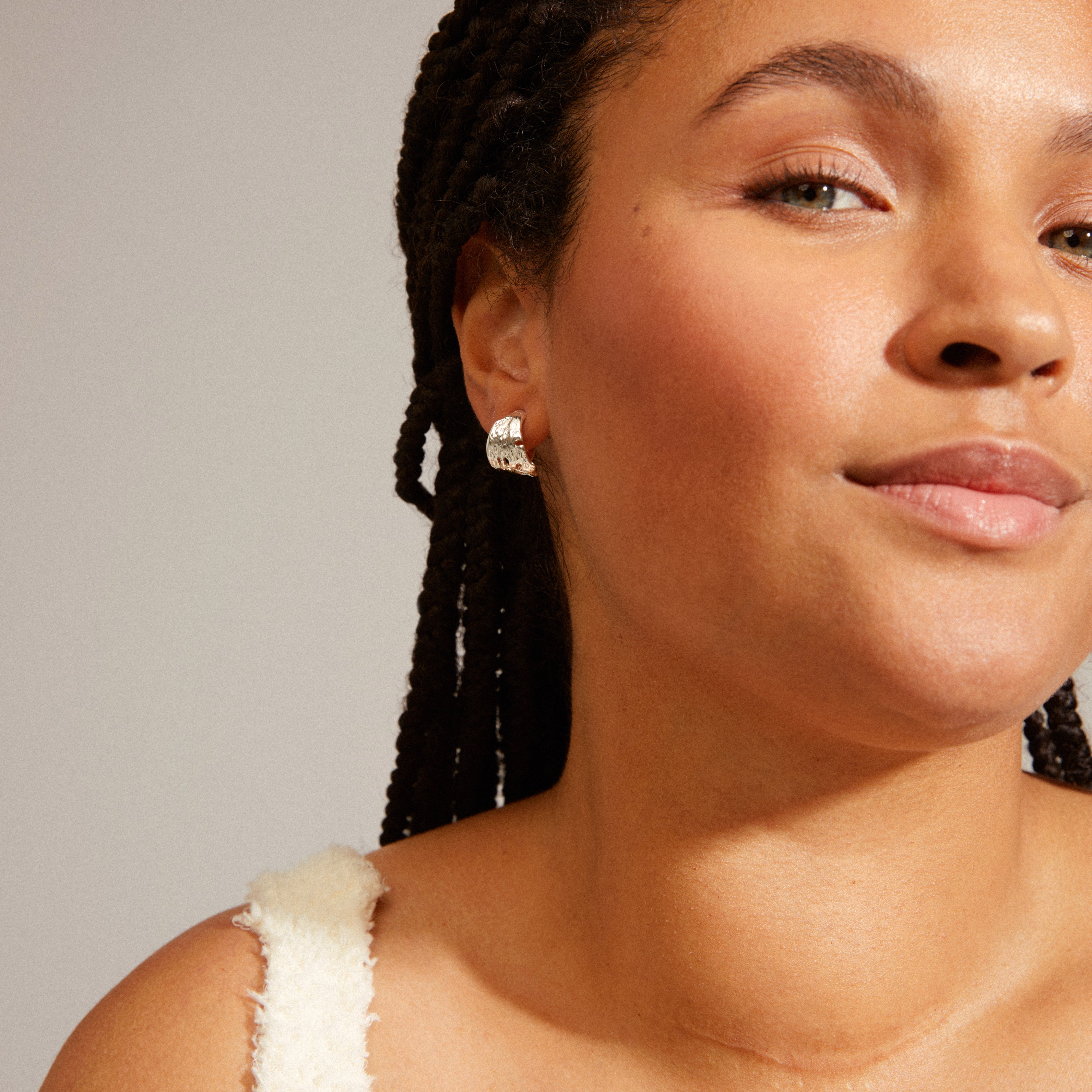 BRENDA earrings silver-plated, featuring wide semi hoops with irregular surfaces, worn by a smiling woman with braids, showcasing their unique handcrafted feel.
