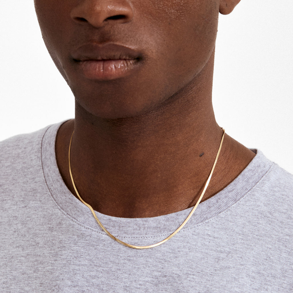 NIKITA necklace gold-plated: A sleek, flat chain elegantly draping above a light gray T-shirt collar, showcasing Pilgrim's chic, genderless style with adjustable length.