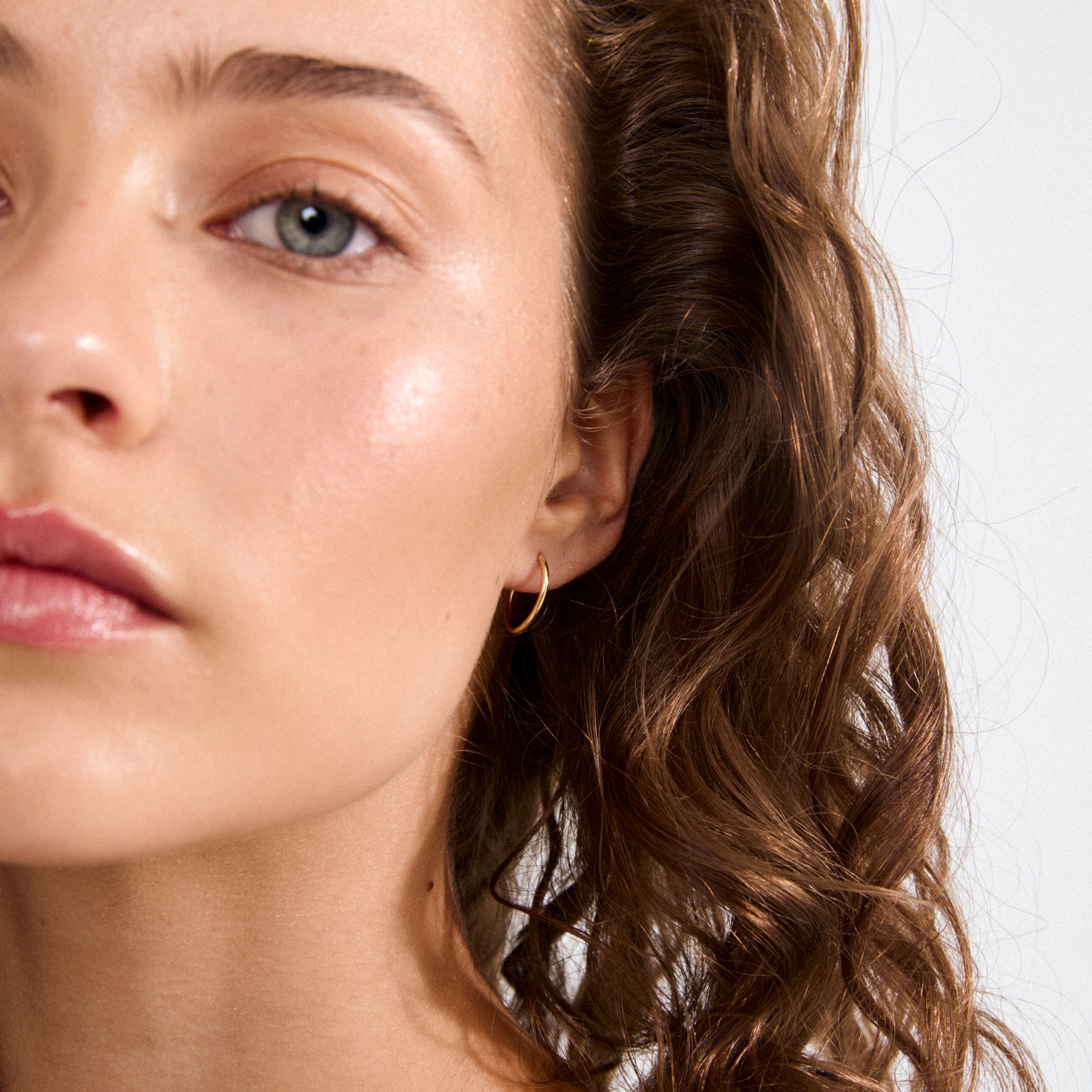 Close-up of a young woman wearing SANNE mini hoop earrings, gold-plated, showcasing her gray-green eye, loose curls, and dewy complexion.