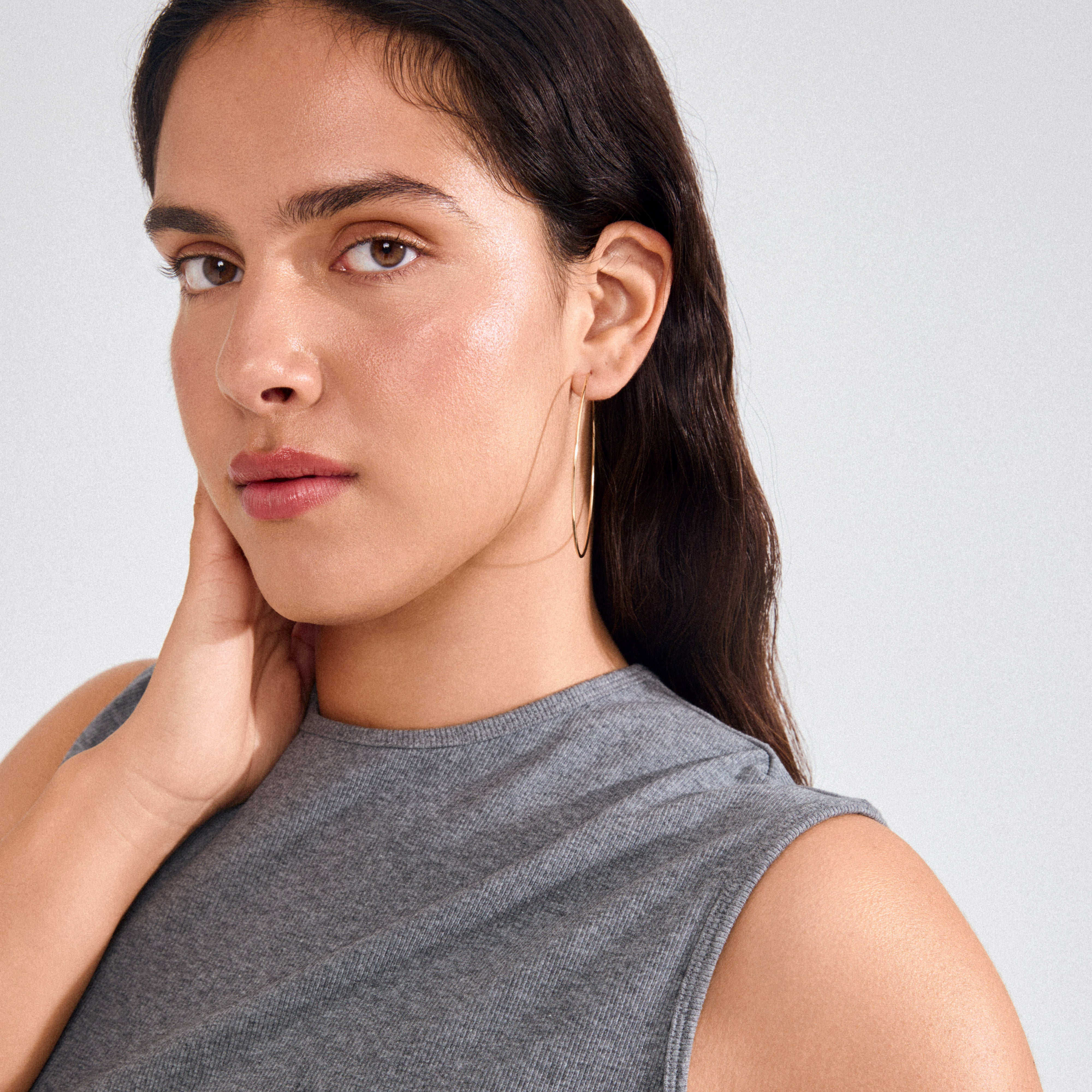 Young woman wearing SANNE X-large gold-plated hoop earring, showcasing its thin, lightweight design. Her hand rests on her neck, highlighting the earring's statement size.