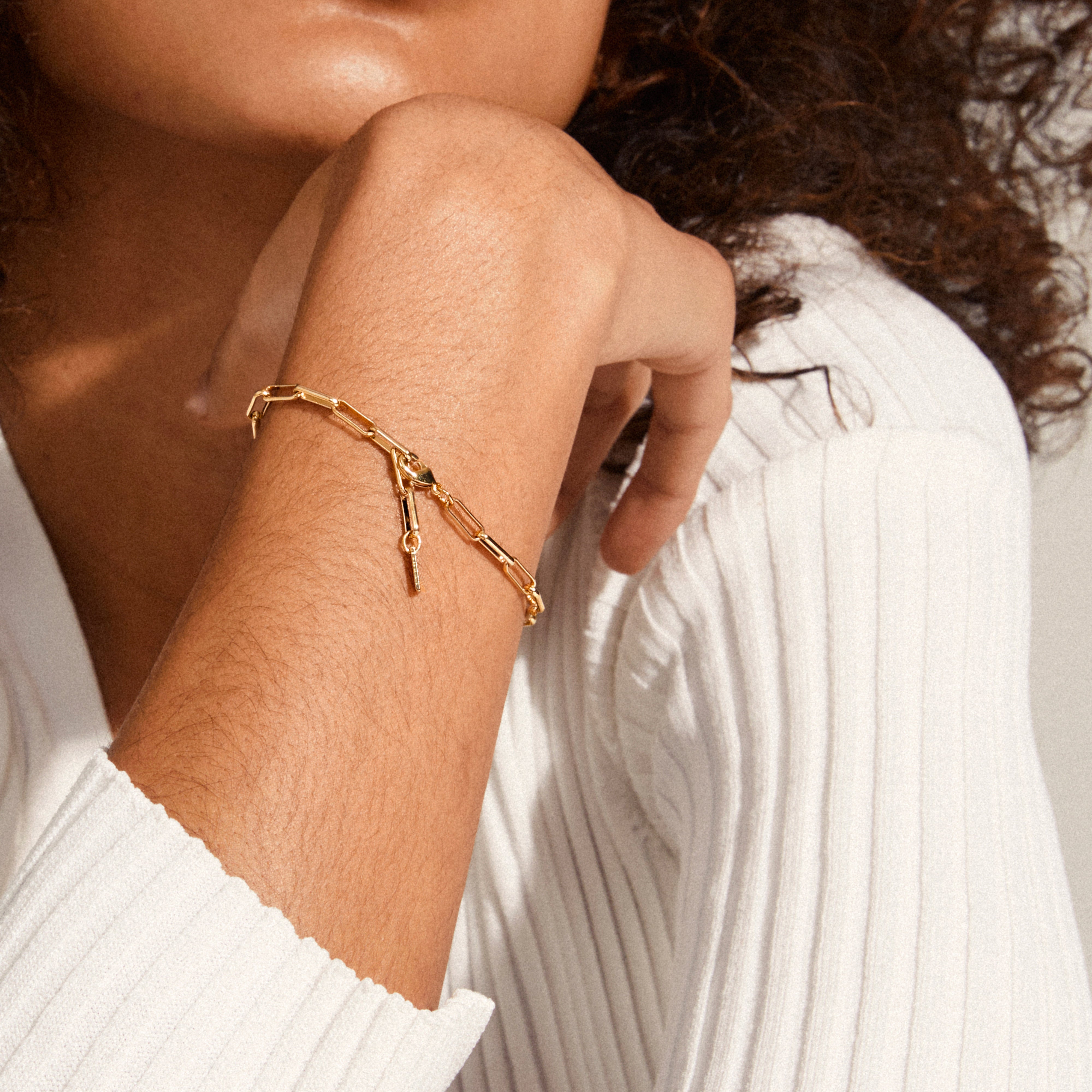 A close-up of a forearm wearing the RONJA bracelet, a gold-plated paperclip chain with a key charm, paired with a ribbed white sleeve.