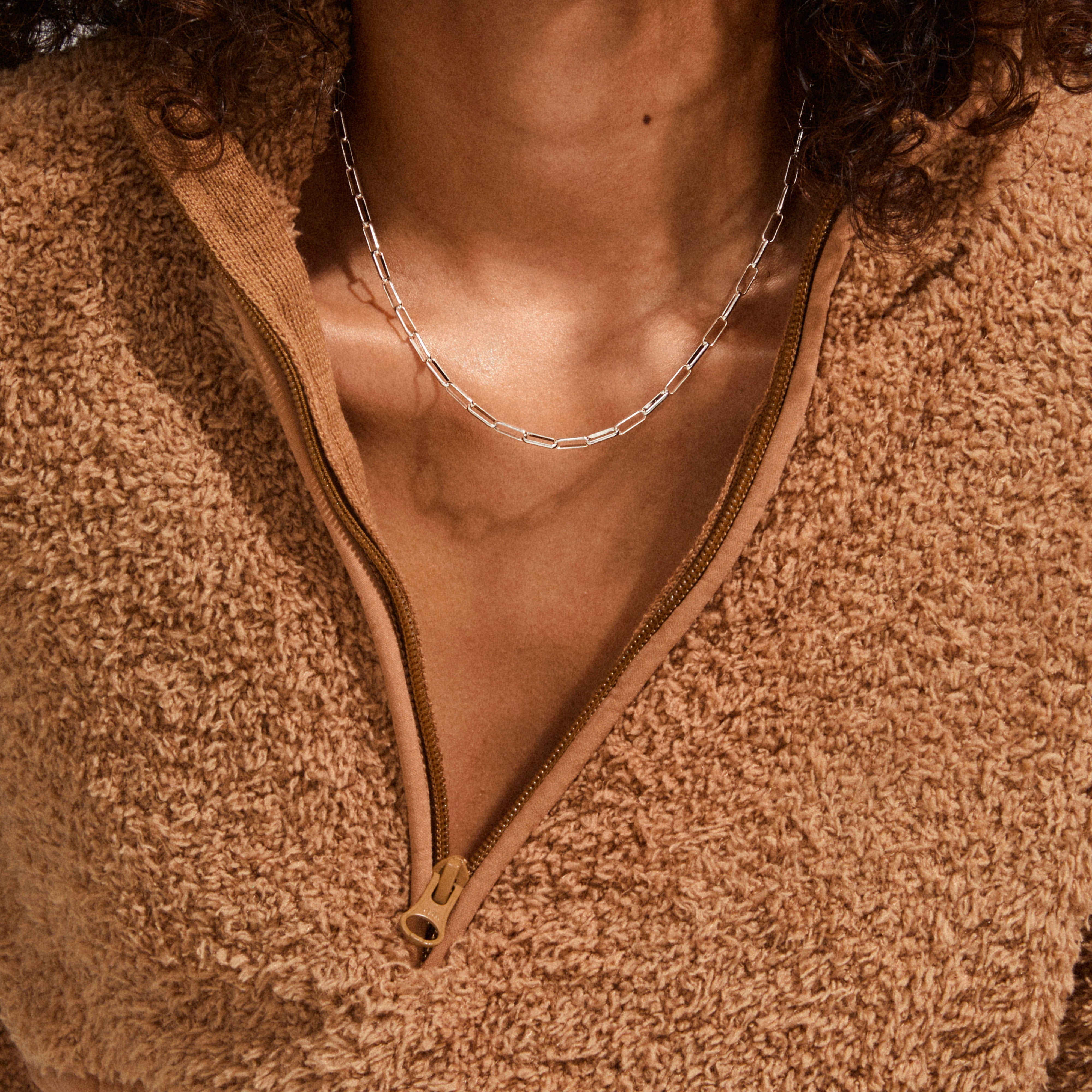 Close-up of a person wearing the RONJA necklace, silver-plated with paperclip links, resting at the collarbone, paired with a tan sherpa-style pullover.