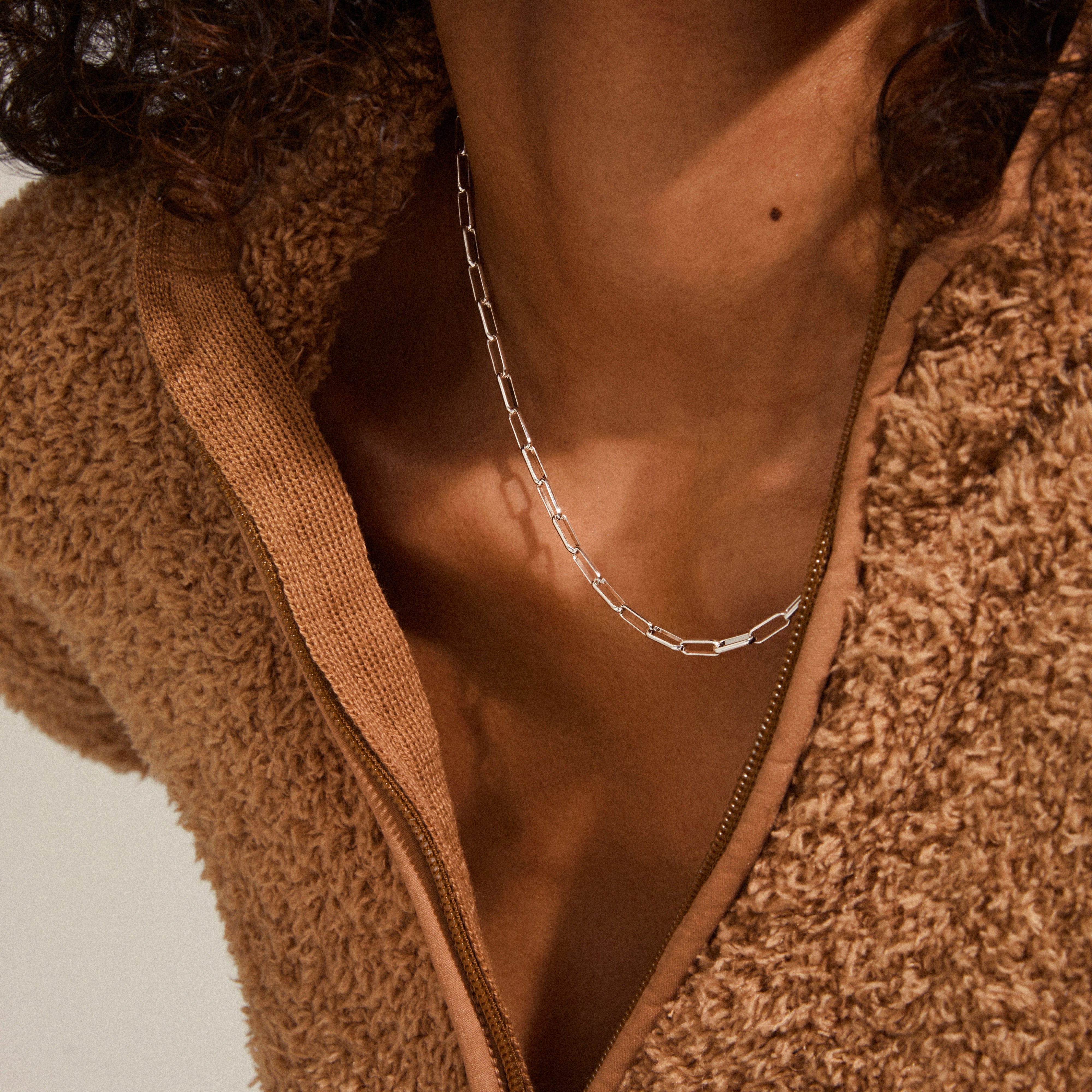 Close-up of a person wearing the RONJA necklace silver-plated, featuring sleek rectangular links around the neck, paired with a partially unzipped teddy-fleece jacket.