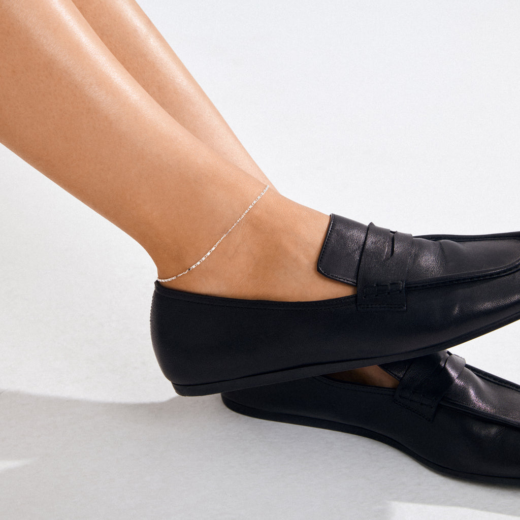 Person wearing sleek black leather loafers with the PARISA flat link anklet, silver-plated, glinting subtly around their ankle.