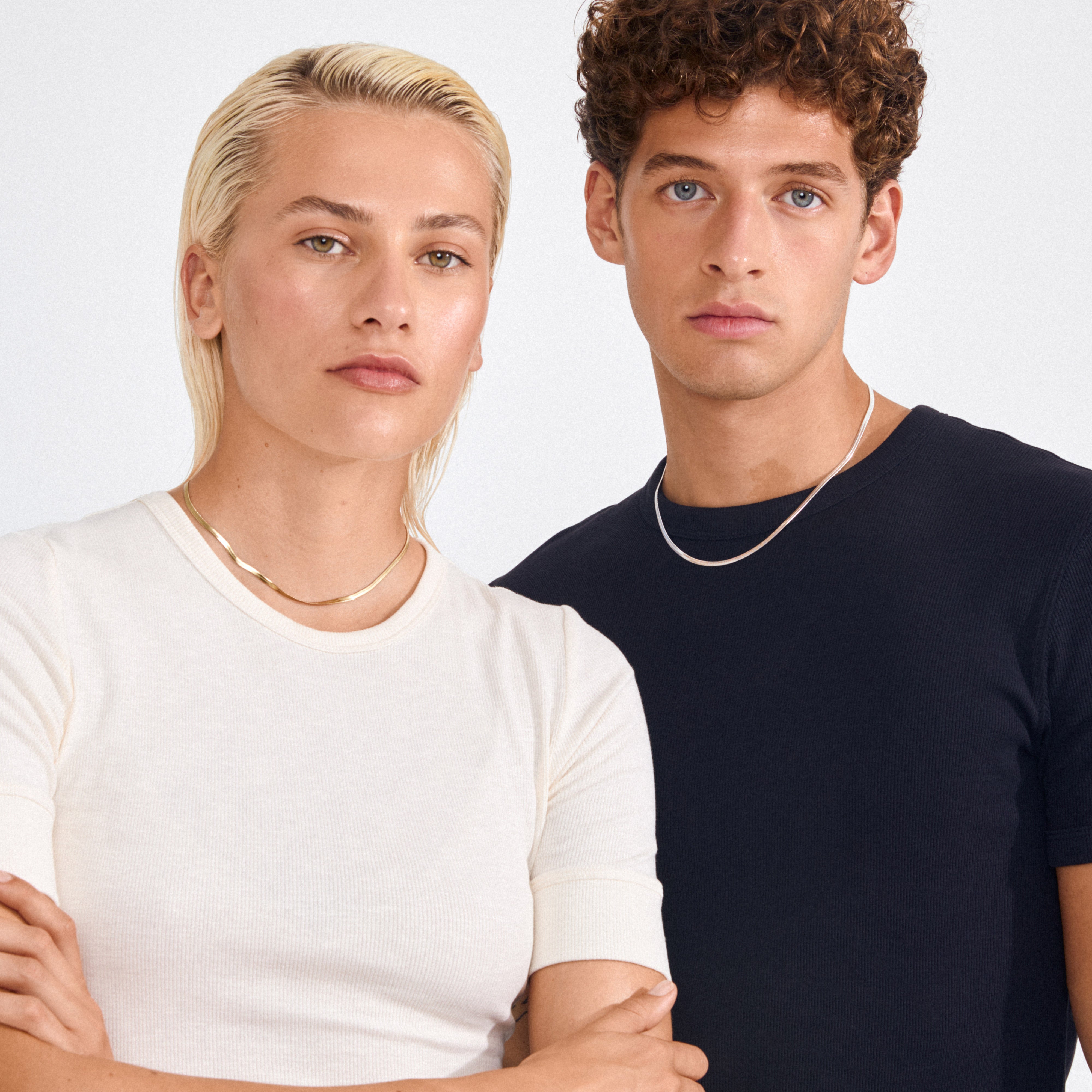Two young adults showcase Pilgrim's JOANNA flat snake chain necklace, silver-plated, styled with casual outfits, highlighting its sleek, versatile design.