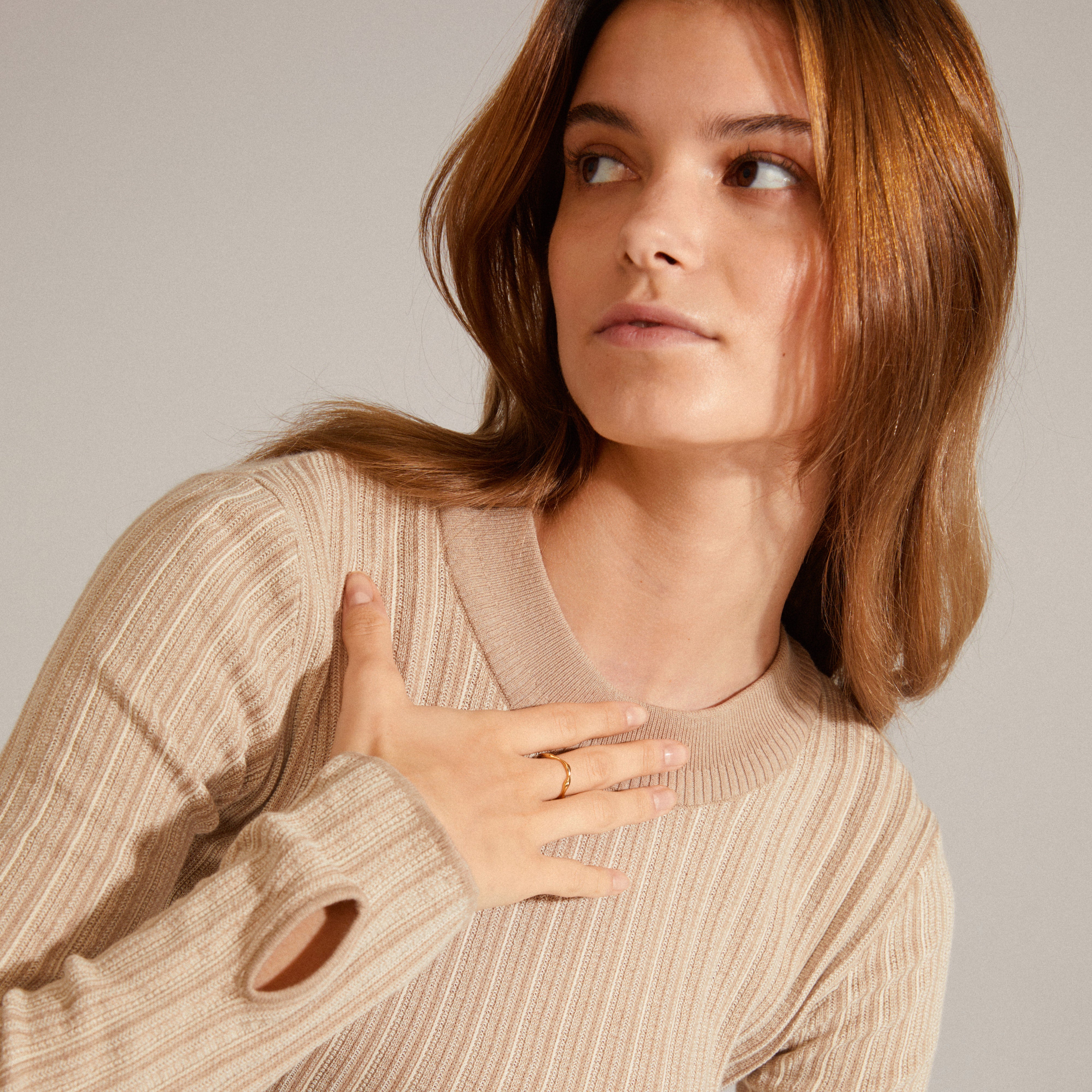 Woman wearing a tan sweater holds her chest, showcasing the LULU organic shaped stack ring gold-plated from Pilgrim, with a handcrafted, wave-inspired design.