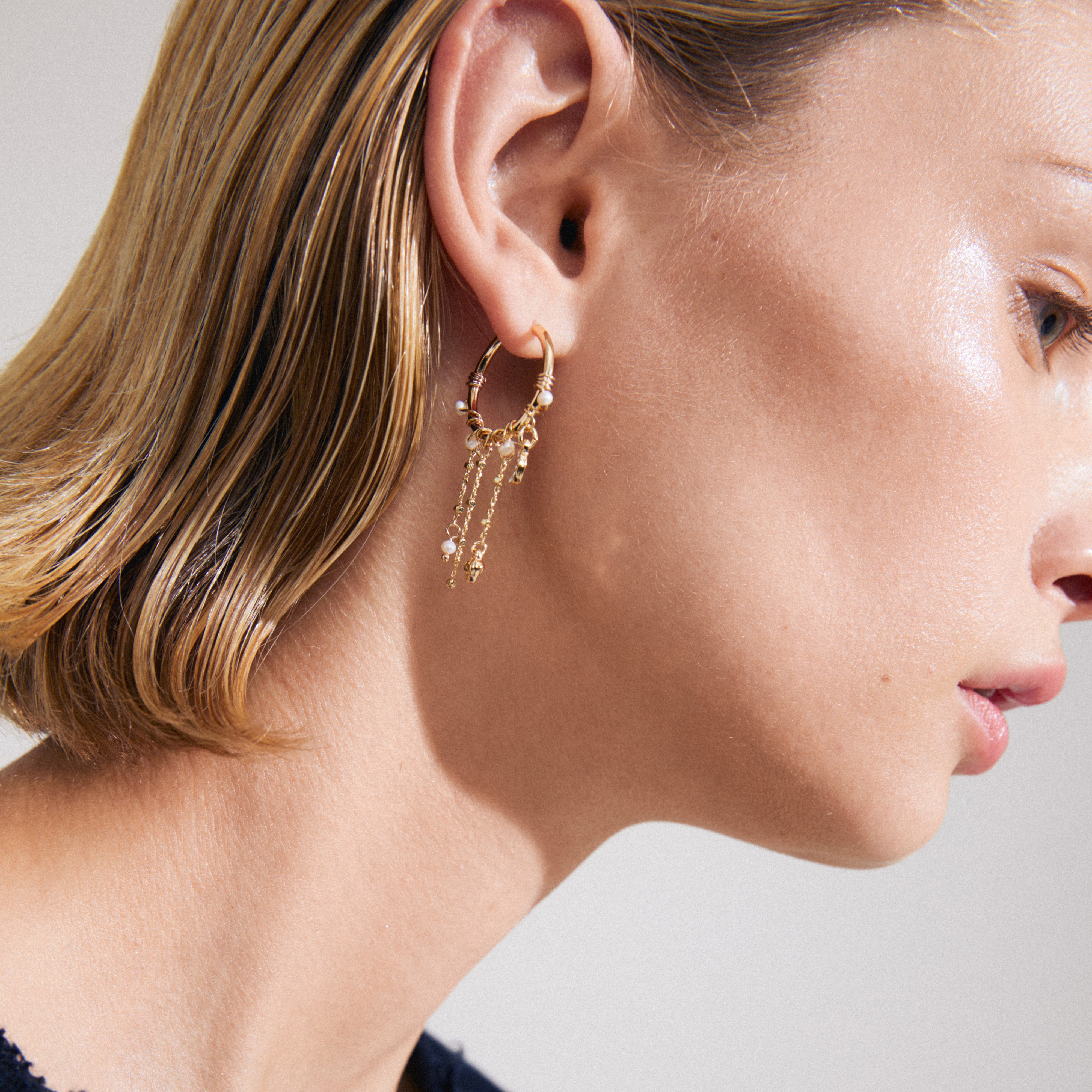 Close-up of a woman wearing ARLET gold-plated hoop earrings with freshwater pearl and Preciosa crystals, featuring tiny moon pendants and hearts.