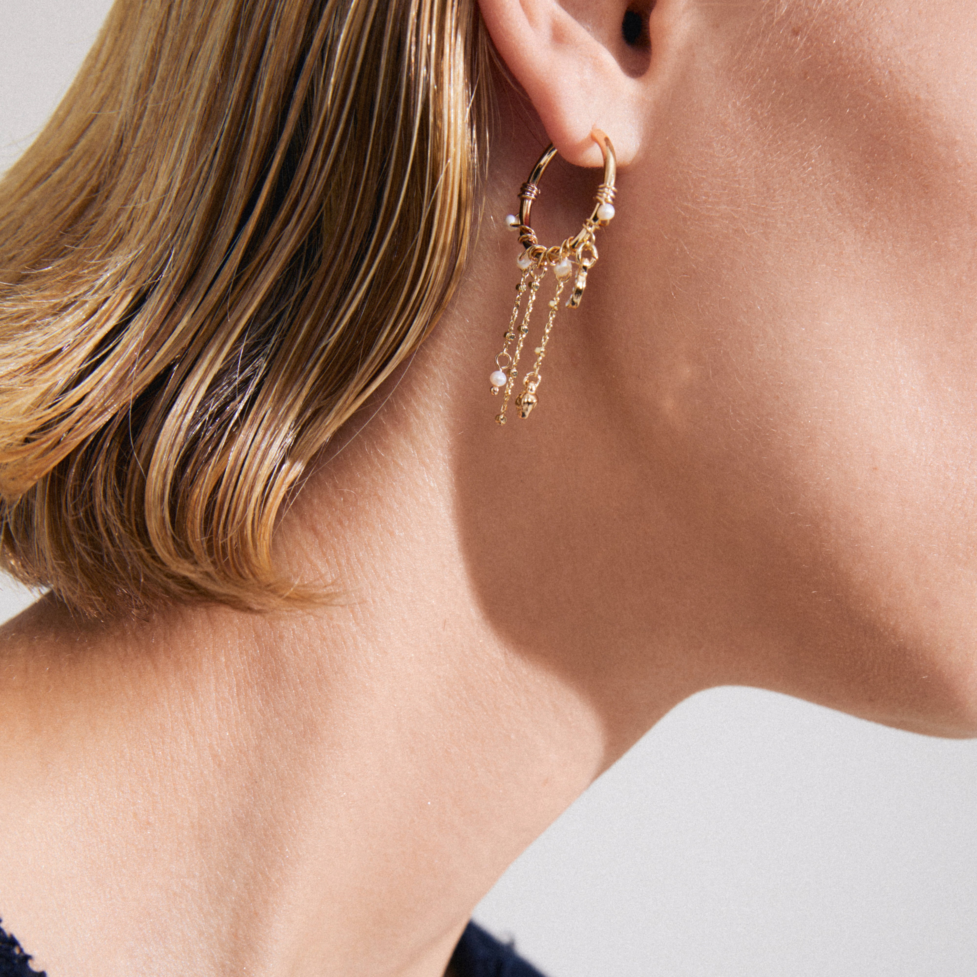 Gold-plated ARLET earrings with dangling chains, pearls, and charms adorn an ear, embodying Pilgrim's boho-lux style and romantic artistry.