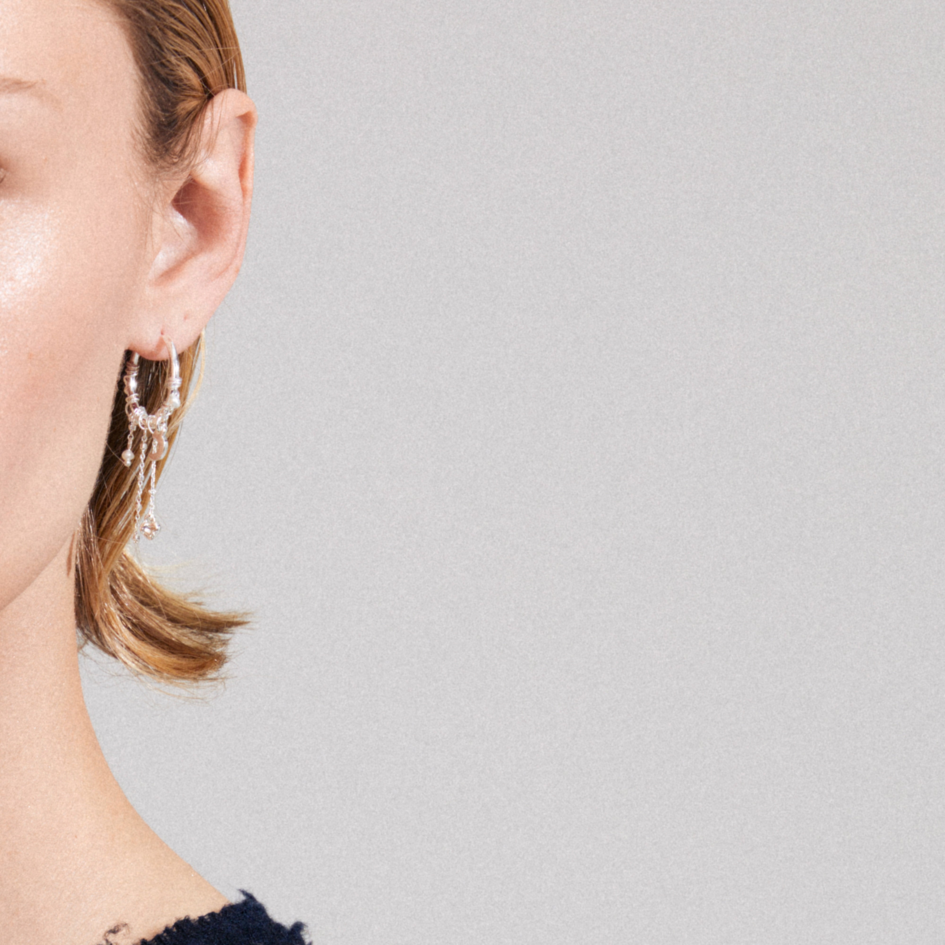 Silver-plated ARLET hoop earring with dangling chains, charms, freshwater pearl, and Preciosa crystals on fair-skinned ear, showcasing artistic, boho-lux style.