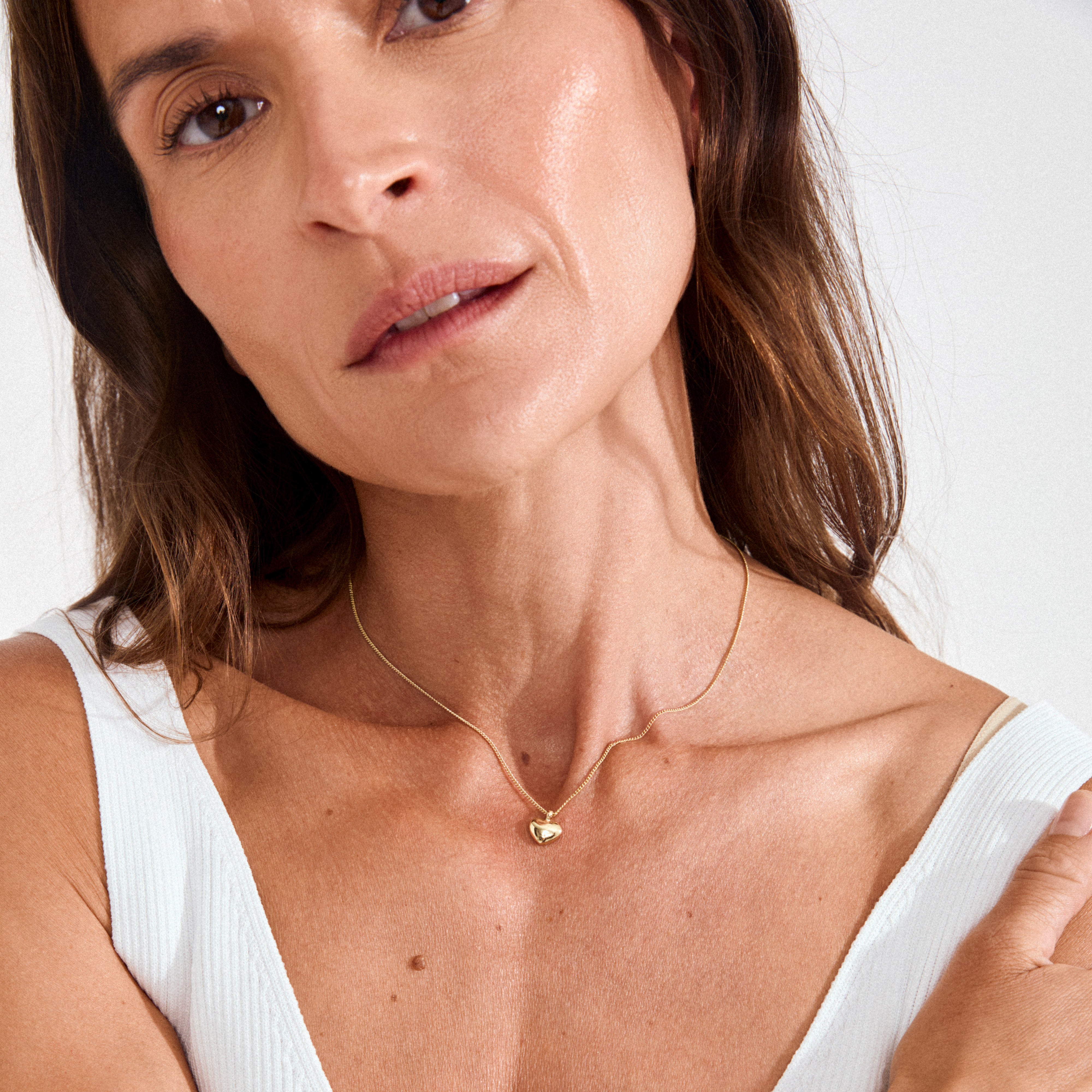 Woman wearing Pilgrim's ARIN gold-plated necklace; a sleek gold chain with a heart pendant, styled over a white ribbed tank top.