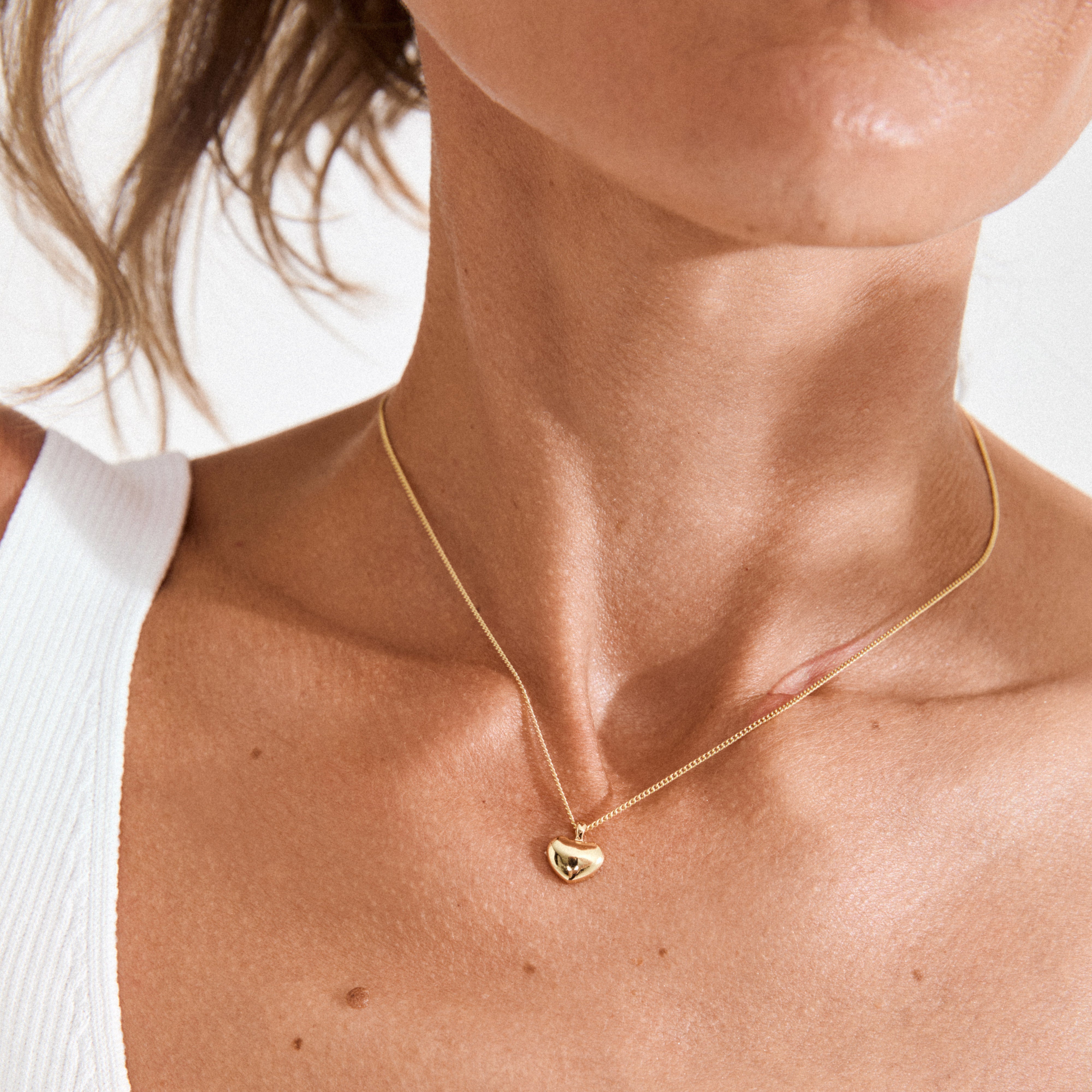 Close-up of the ARIN necklace gold-plated, featuring a sleek heart pendant on a woman's neck, styled with a white ribbed tank, highlighting its subtle elegance.