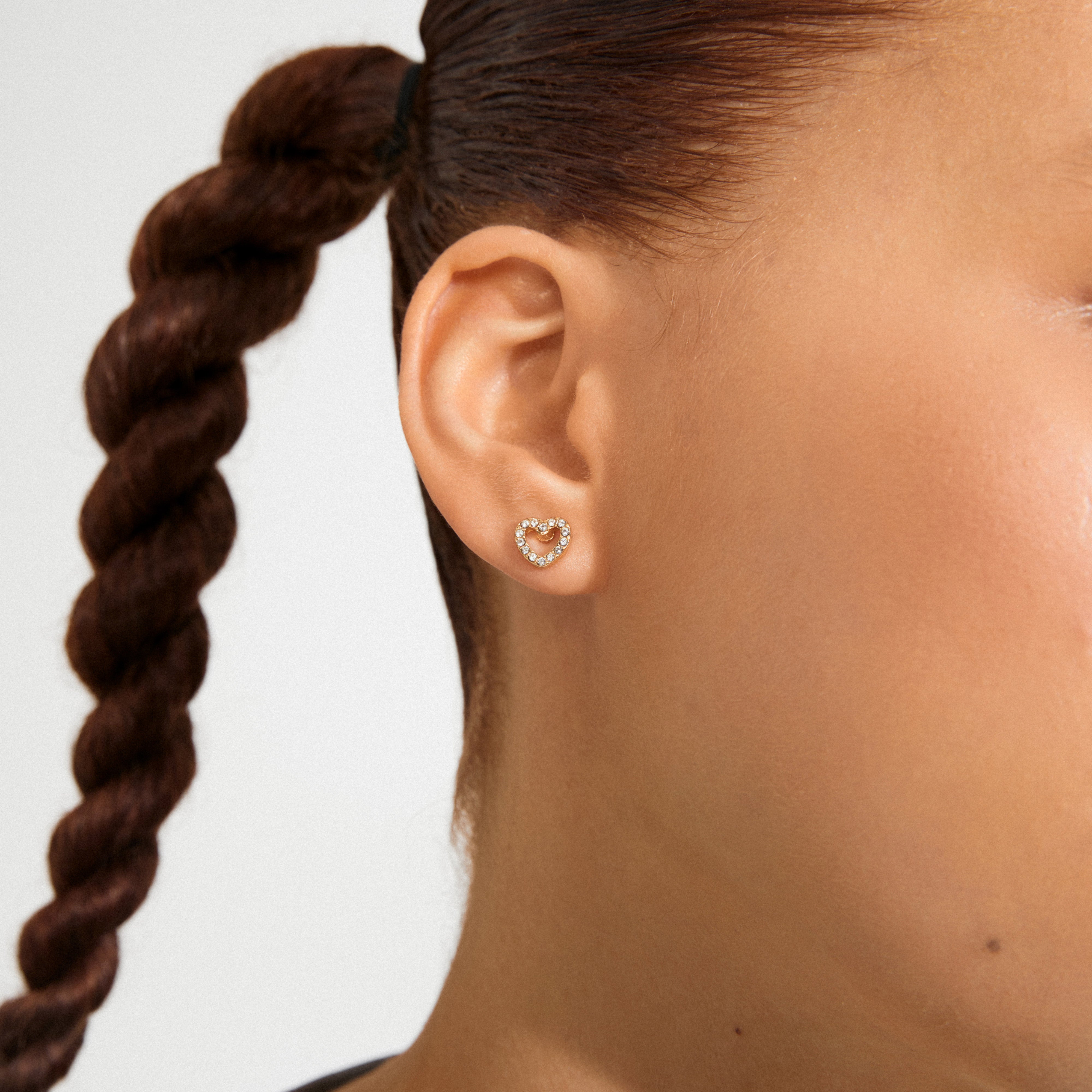 EDIE crystal heart earrings gold-plated, encrusted with sparkling stones, adorn a person's earlobe, offering a chic and romantic touch to any outfit.