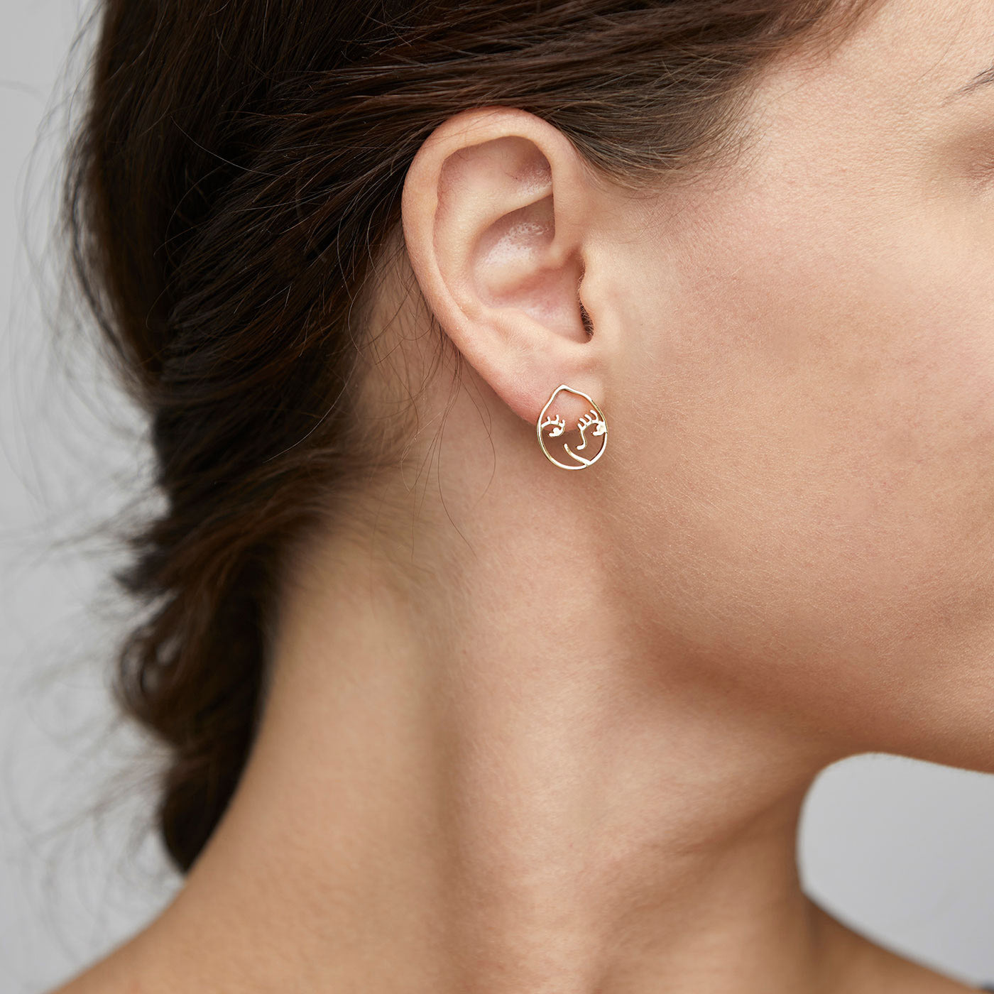 Close-up of DEBRA earrings, gold-plated with an artistic smiling face design, worn on a woman's ear, showcasing unique style and creativity.