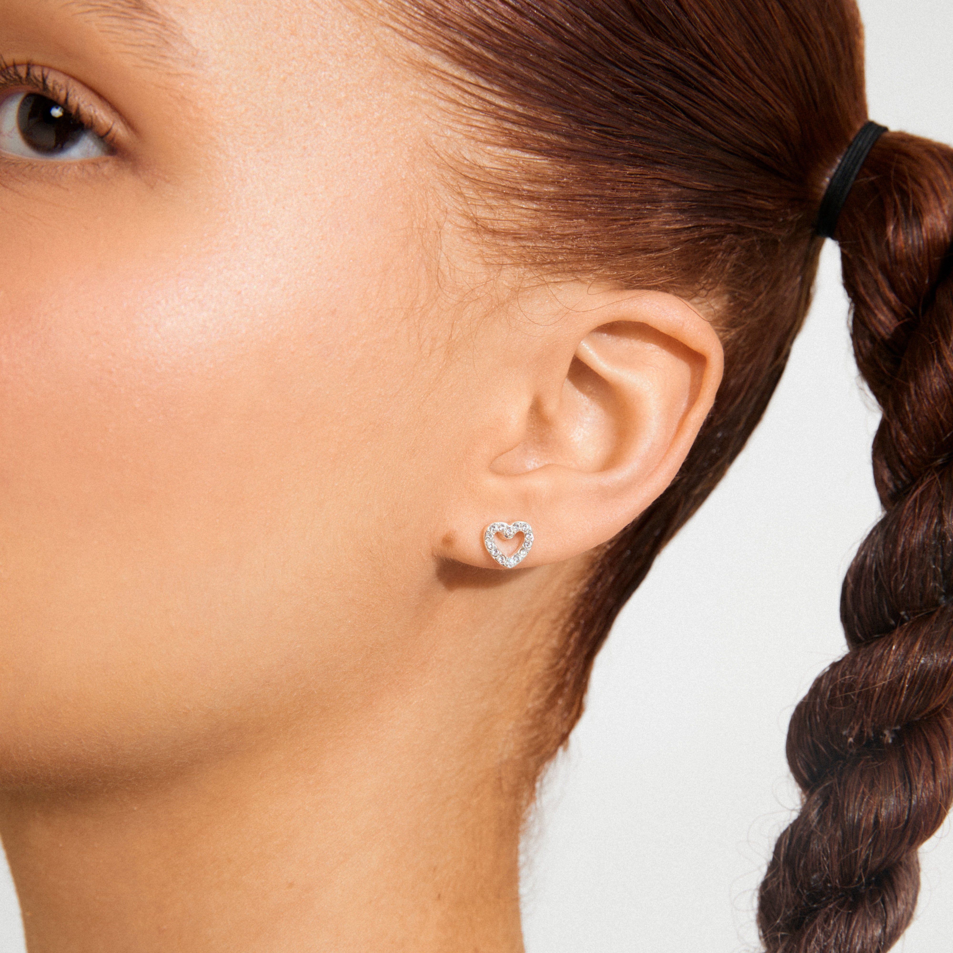 EDIE crystal heart earrings, silver-plated, sparkle with crystals on a person’s ear, complementing a sleek braided hairstyle, showcasing their delicate, romantic charm.