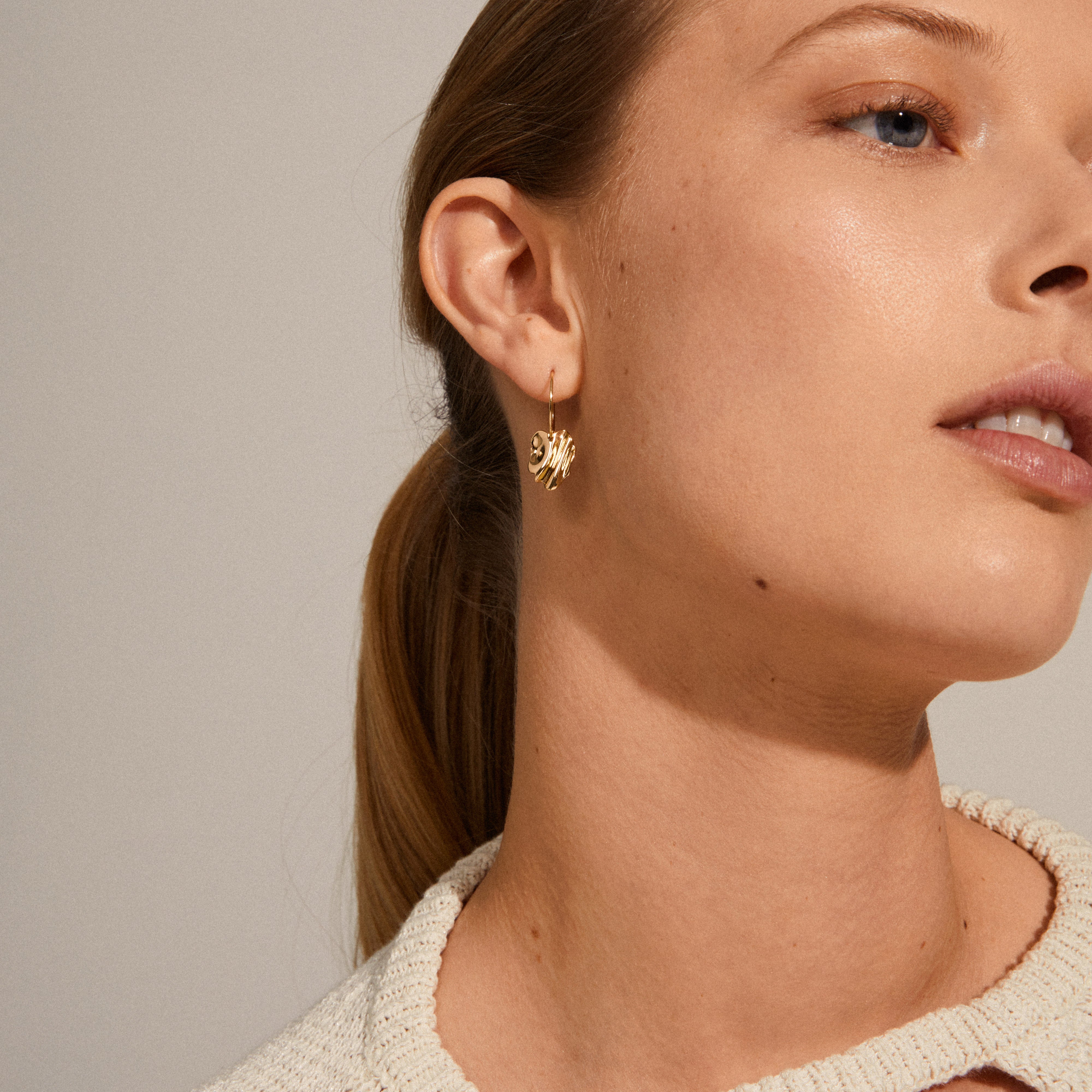 Close-up of a woman wearing Pilgrim's EM wavy hoop earrings, gold-plated, showcasing nature-inspired, asymmetrical design with a handcrafted feel.