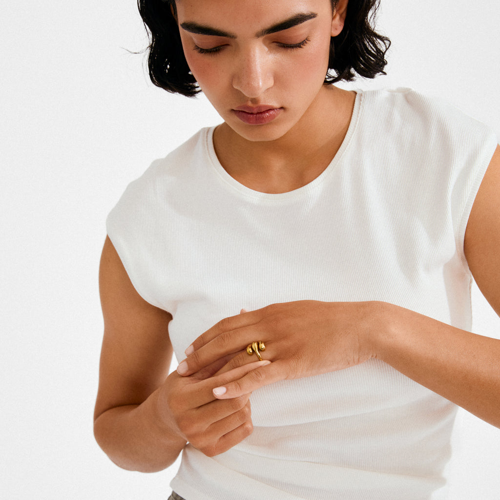 Young person wearing a white top highlights the gold-plated JENNINGS ring by Pilgrim, showcasing its sculptural, melting design on their finger.