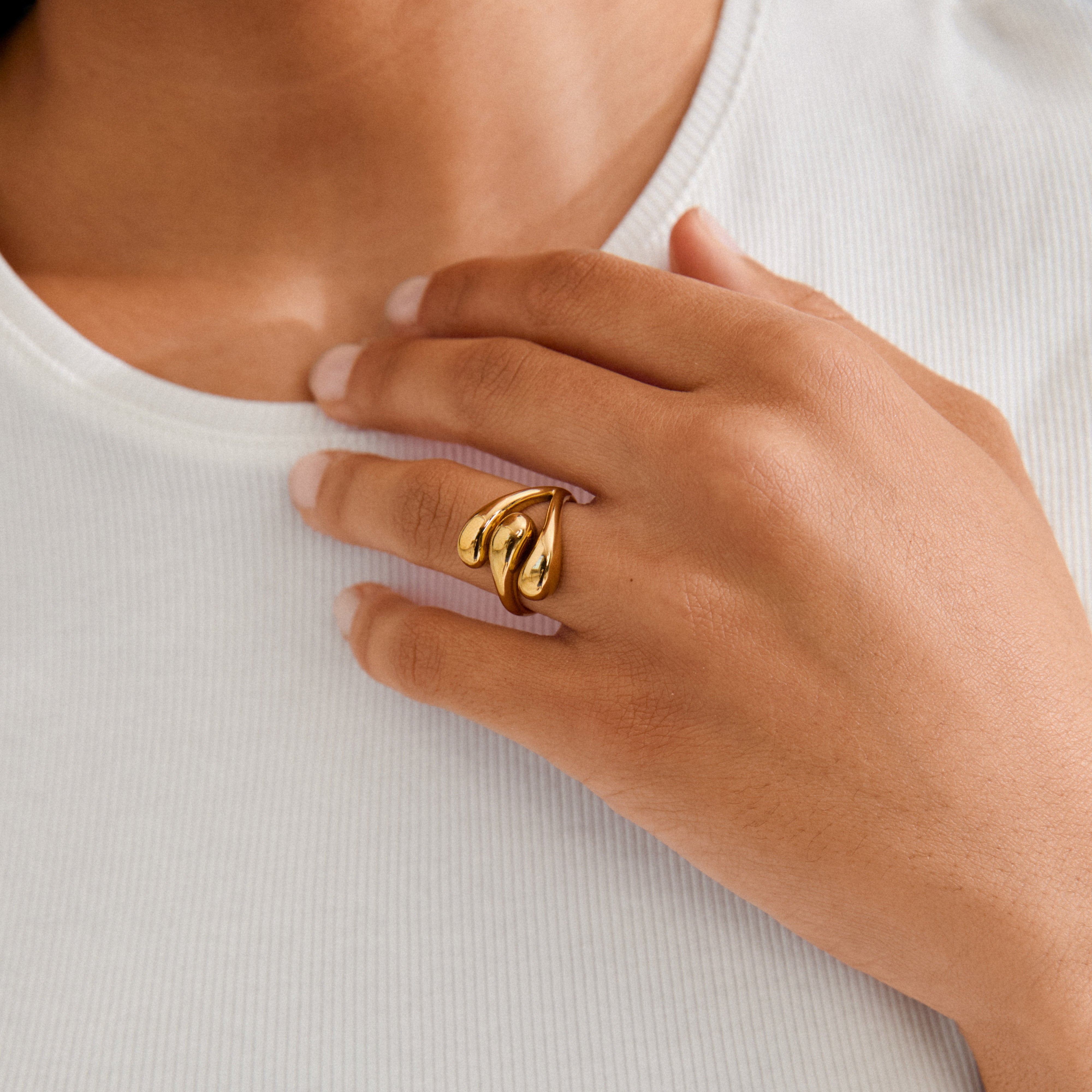 A hand wearing the gold-plated JENNINGS ring by Pilgrim, featuring three sculptural, droplet-like forms, resting on a rib-knit top.