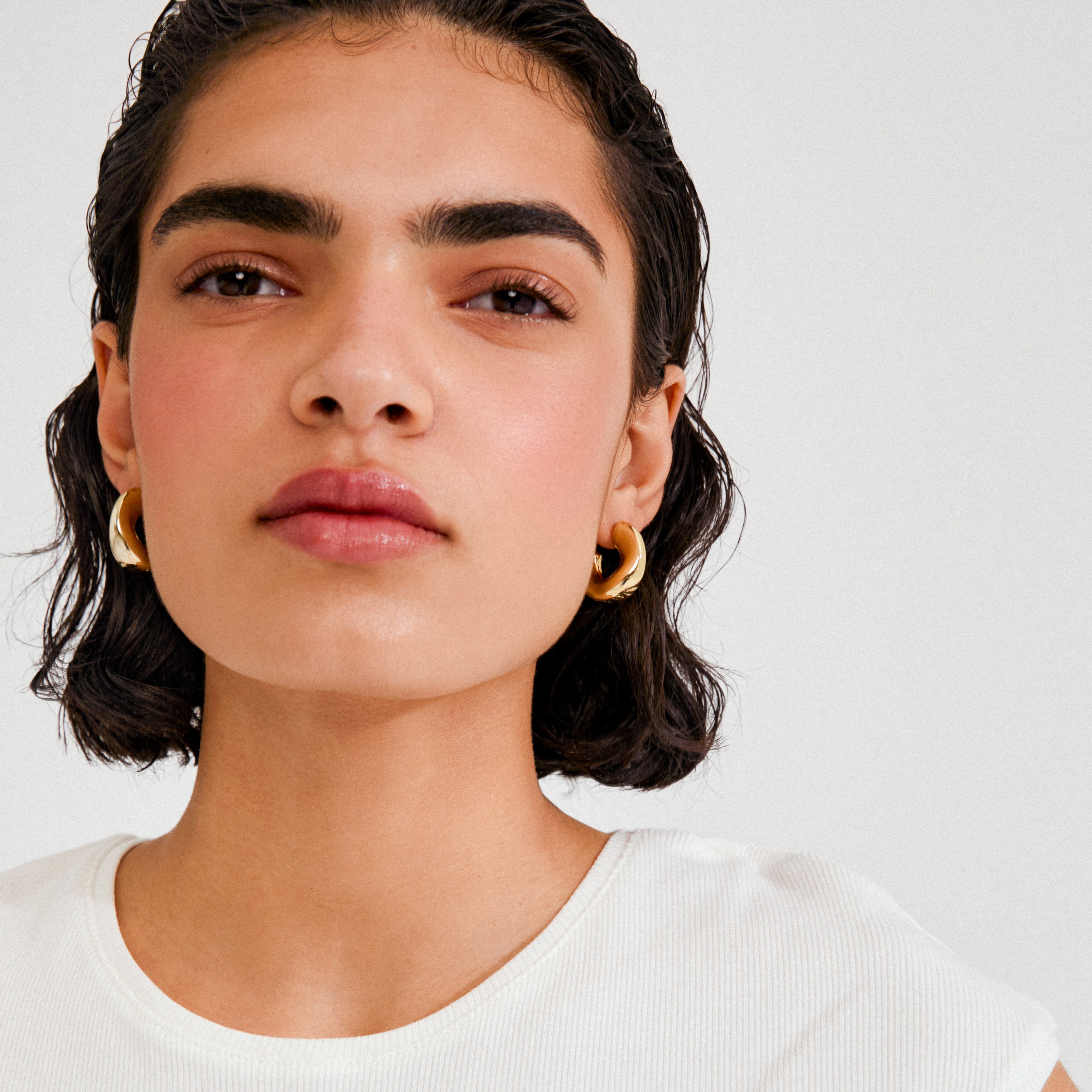 Young woman wearing JENNINGS hoop earrings gold-plated by Pilgrim, featuring chunky, sculptural gold hoops that hug the ear, styled with short, dark hair.