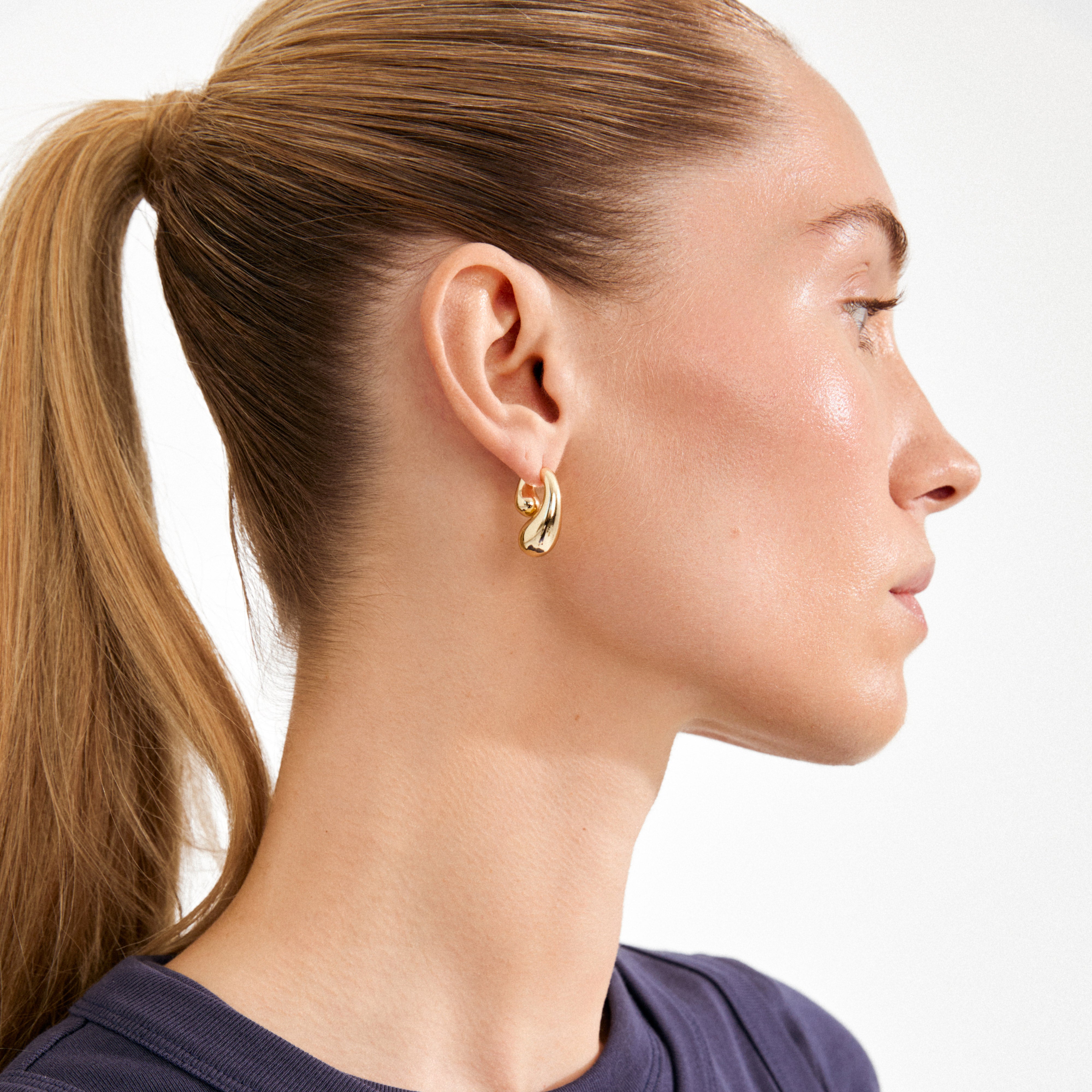 Close-up of a young woman wearing Pilgrim's gold-plated JENNINGS earrings, featuring a sculptural, double melting-drop design that wraps around the ear, highlighting its 3D effect.