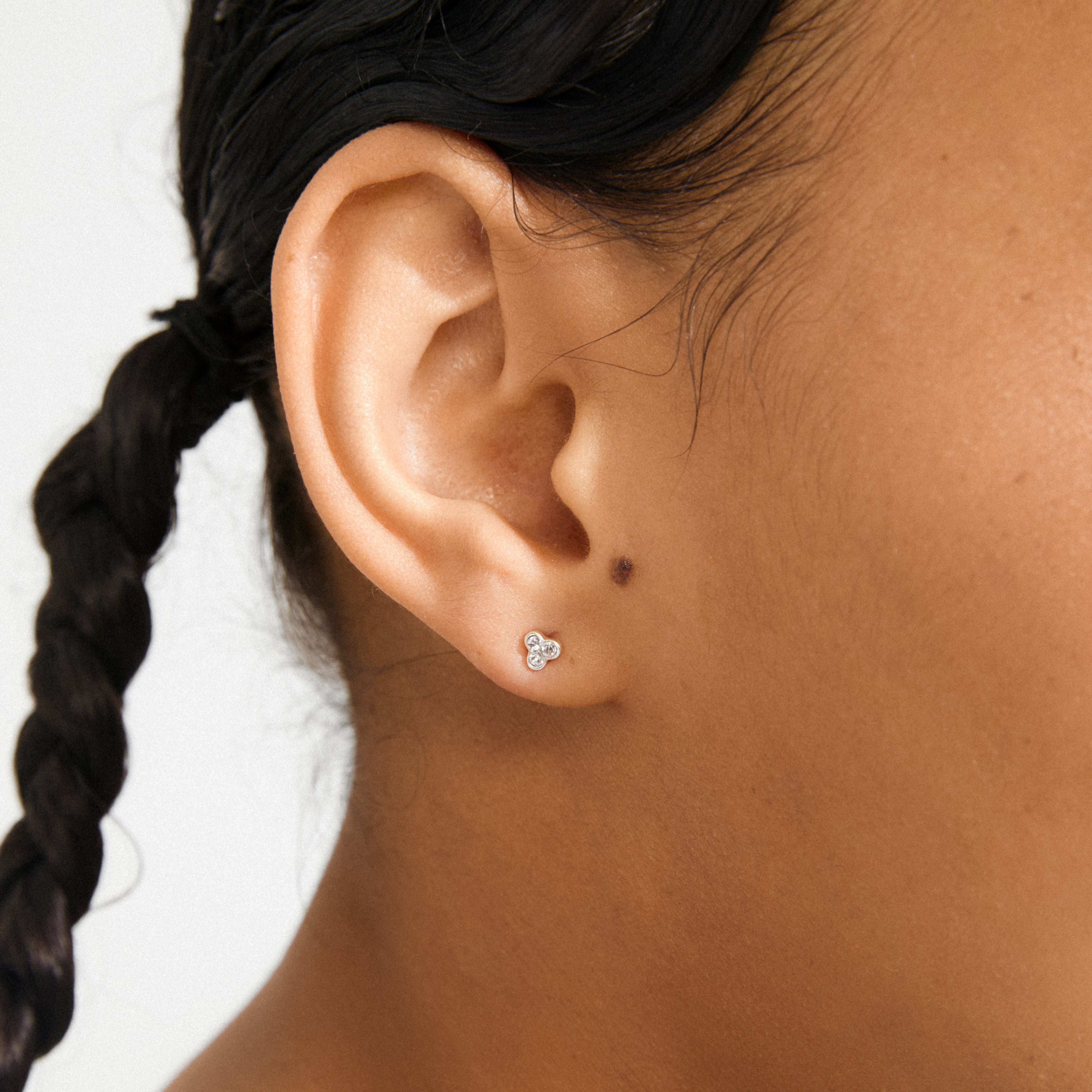 CAILY crystal earrings, silver-plated, featuring a trio of clear gemstones in a clover-like cluster, adorn a medium tan ear with a dark braid behind.