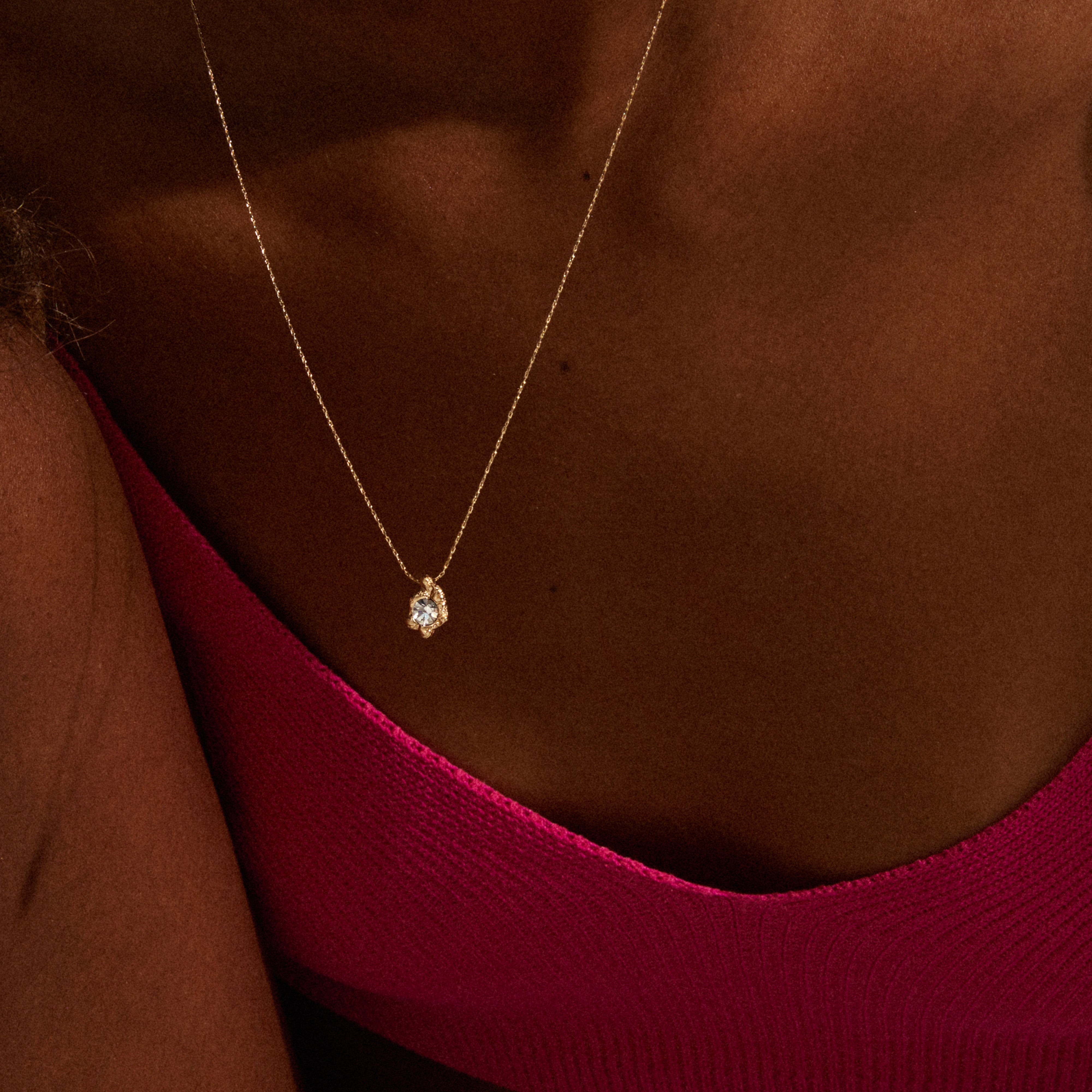 TINA crystal pendant necklace gold-plated on thin chain, featuring a clear gemstone on a person's décolletage, worn with a ribbed pink top.