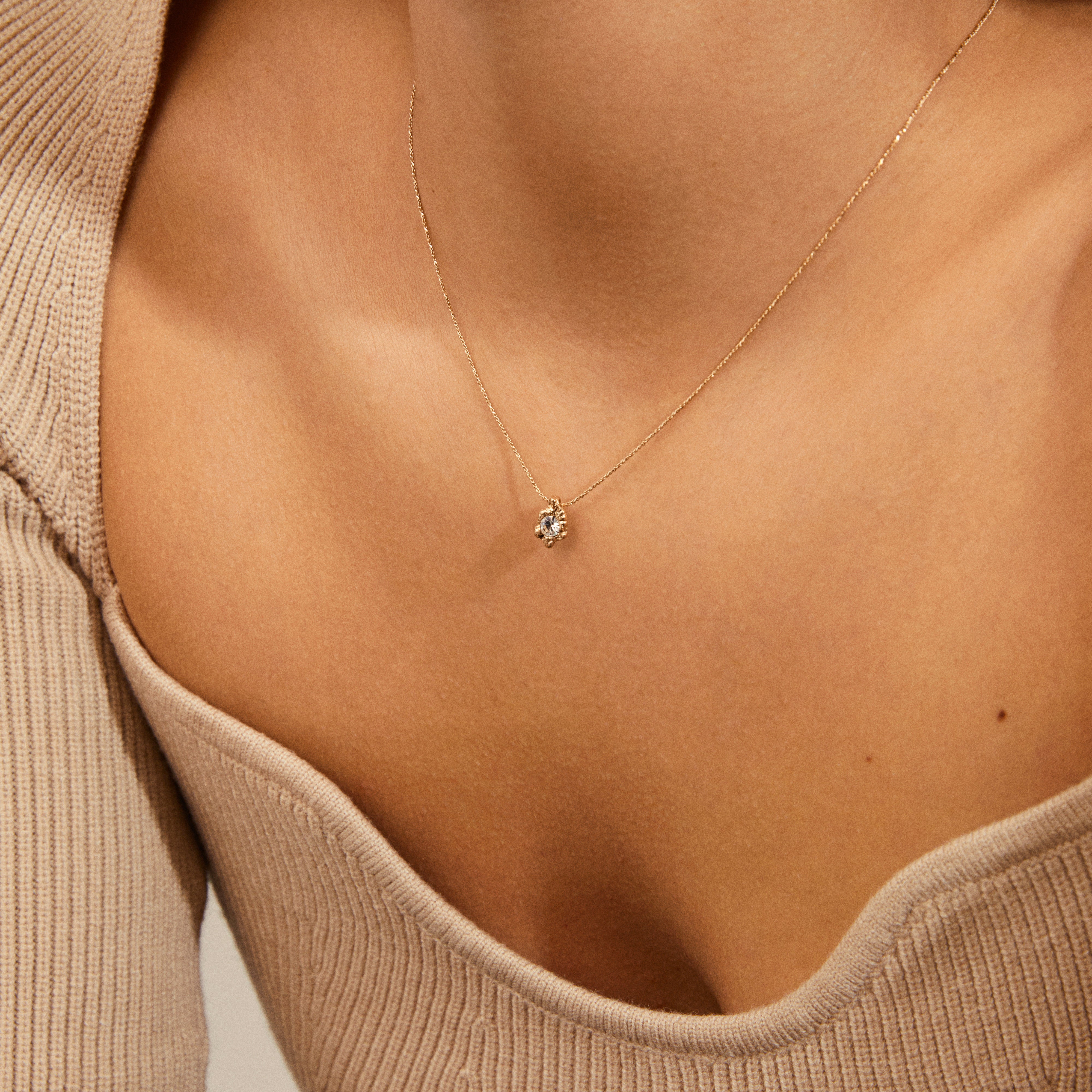 Close-up of TINA crystal pendant necklace gold-plated by Pilgrim, featuring a fine gold chain with a clear gemstone pendant against a ribbed knit top.
