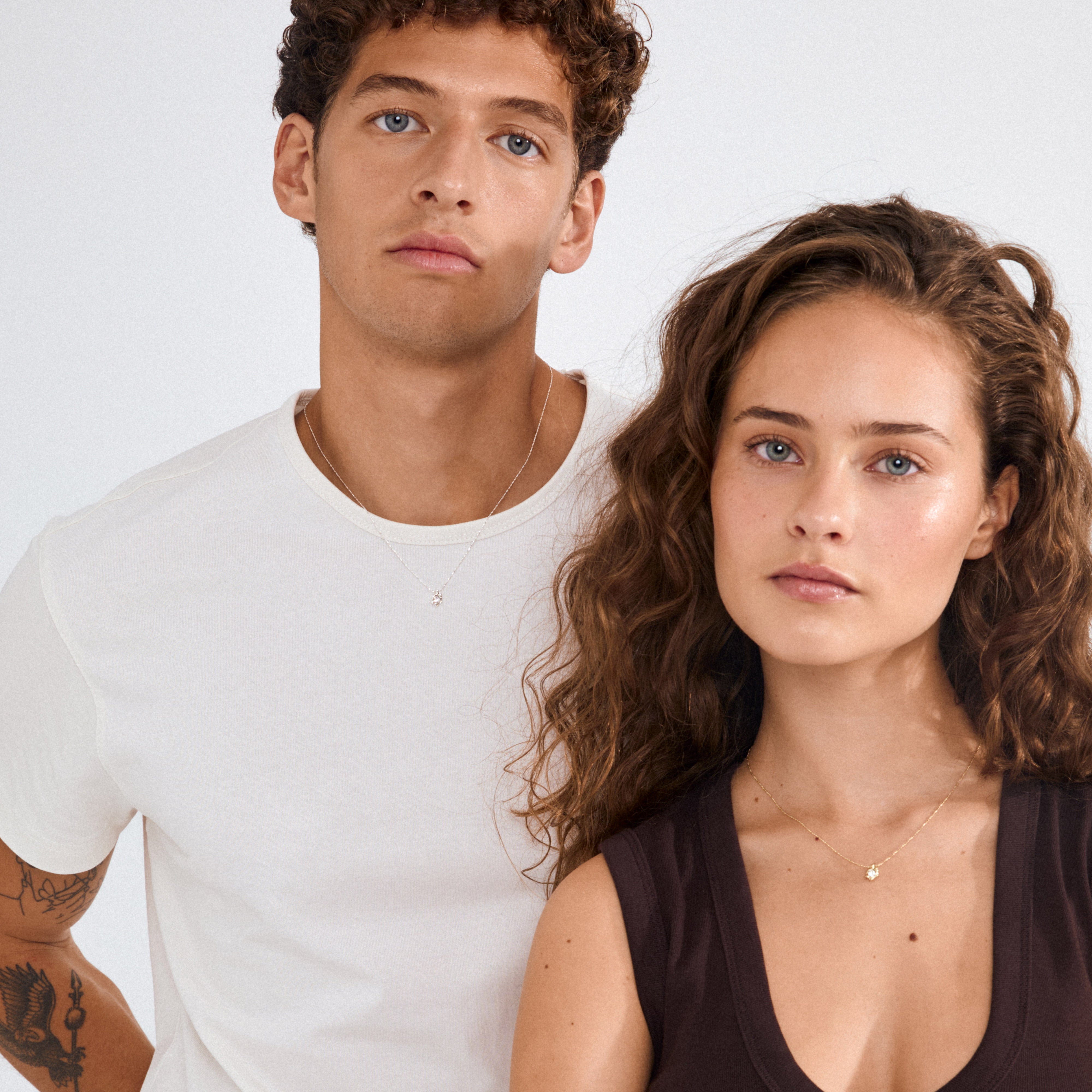 Young man and woman wearing the TINA crystal pendant necklace, showcasing its silver-plated, sculptural design with a crystal charm against neutral studio backdrop.