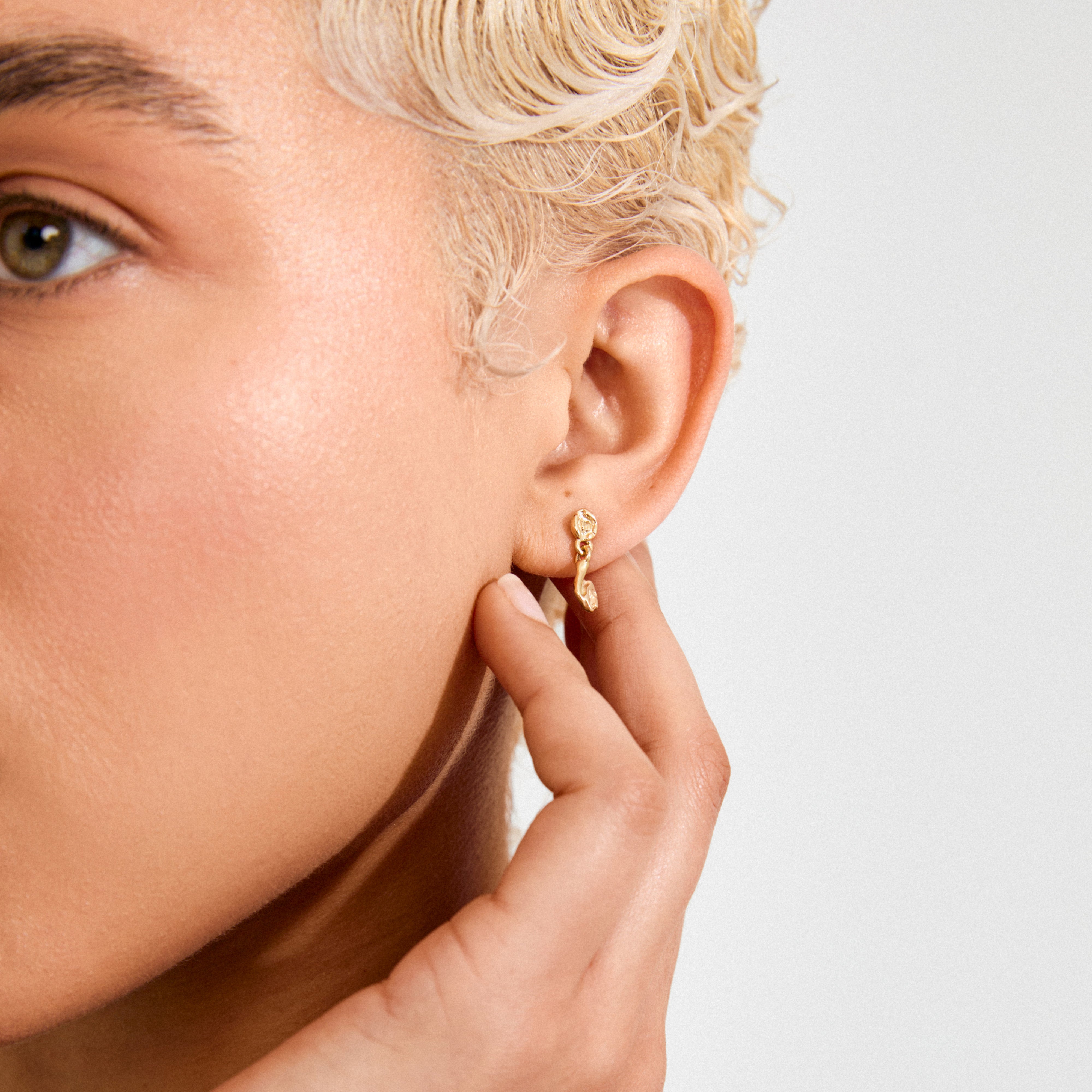Close-up of a person's ear wearing the gold-plated ELIYAH earrings from Pilgrim, featuring a textured, irregular-shaped pendant gently touched by a hand.