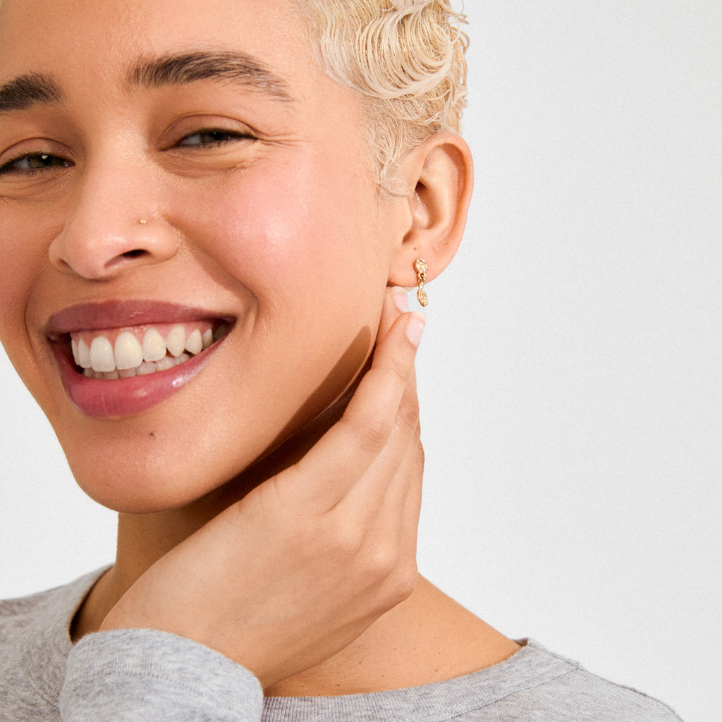 Person wearing ELIYAH gold-plated earrings by Pilgrim, showcasing their sculptural, textured design with dangling movement, complemented by a light gray top.