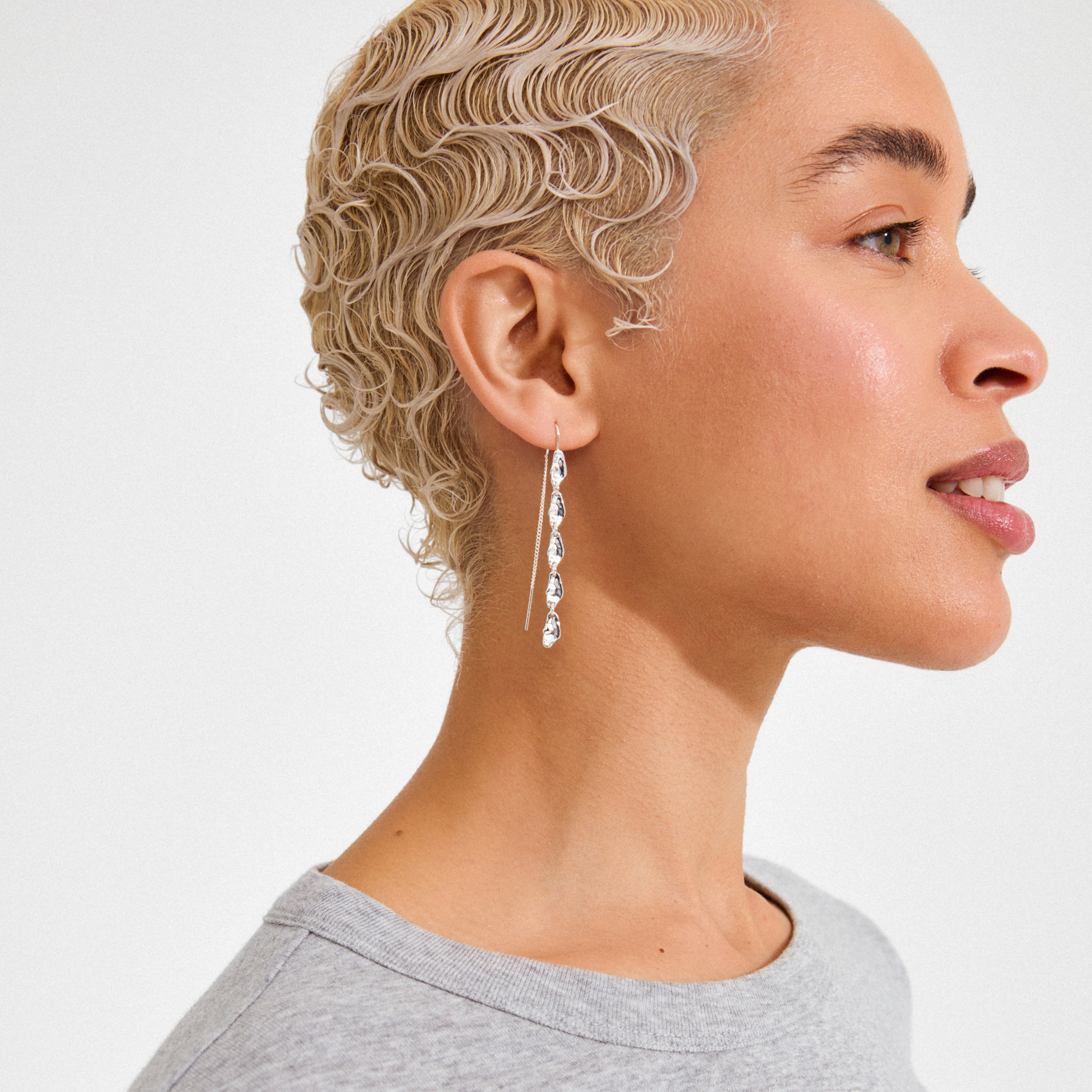 ELIYAH chain earrings silver-plated, featuring sculptural, droplet-shaped charms, hang elegantly, complementing the model's bleached finger-wave hair against a plain white background.
